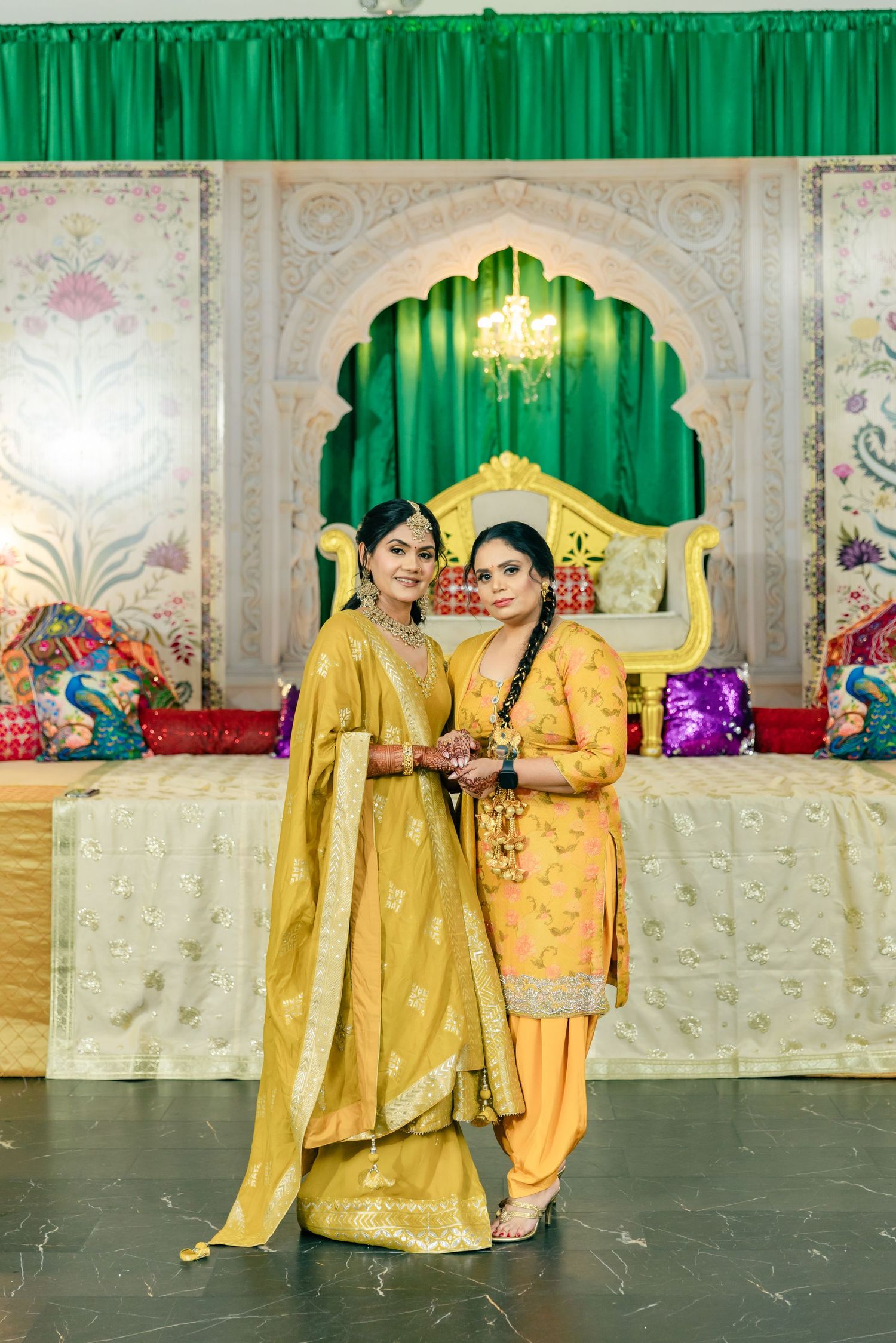 Two women in traditional yellow Punjabi outfits pose together at an elegant wedding venue with green curtains.