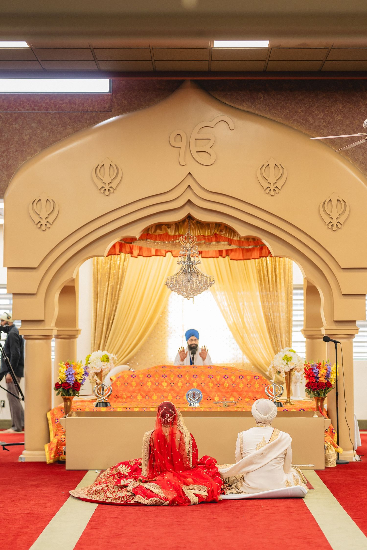 Traditional Sikh wedding ceremony setup with ornate red and gold decorations in a Gurdwara featuring arched backdrops.