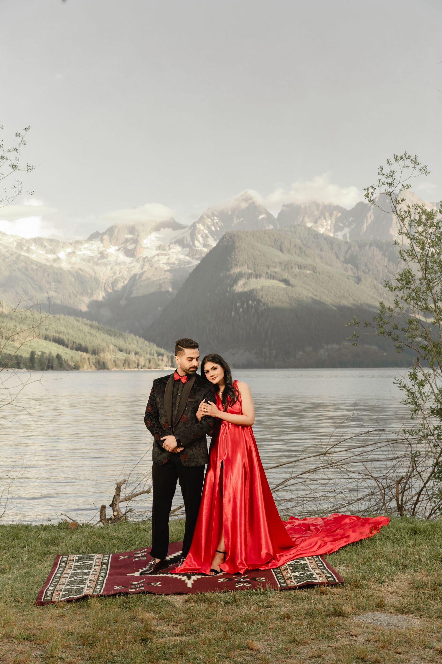 Couple in formal attire posing on red blanket by mountain lake.