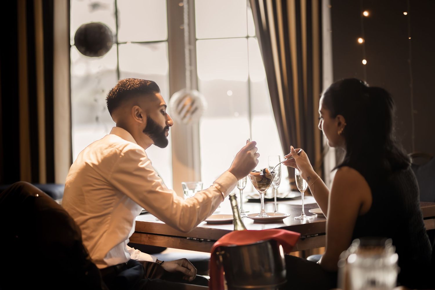 A bearded man and a woman with a ponytail share a romantic dinner at a candlelit restaurant table by a large window.