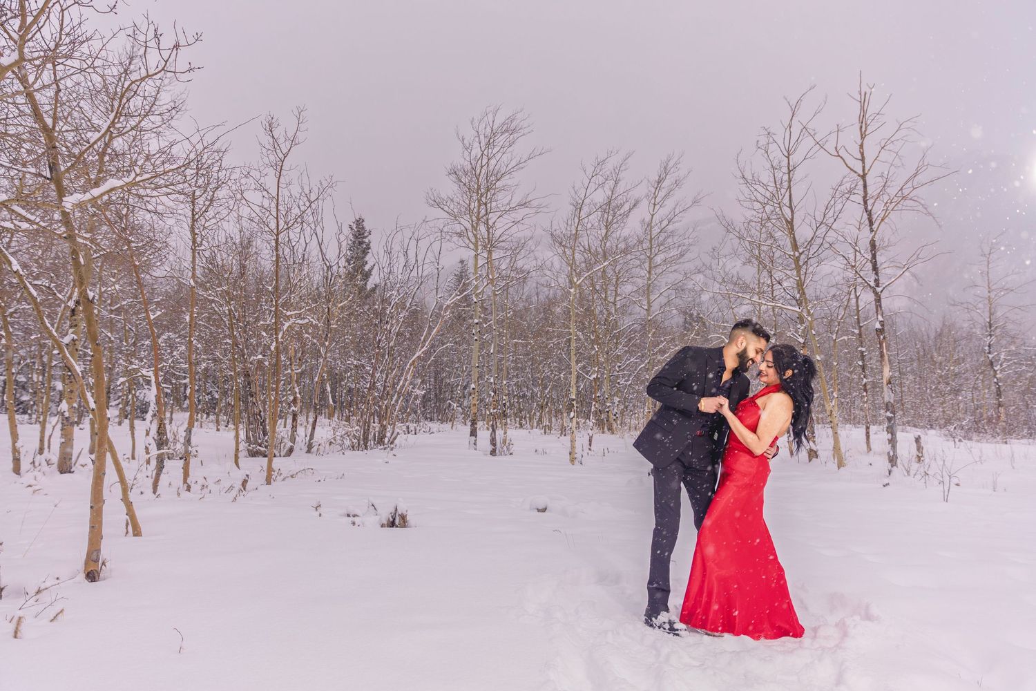 Couple embracing in snowy winter forest, woman in red gown and man in dark suit, surrounded by bare trees.