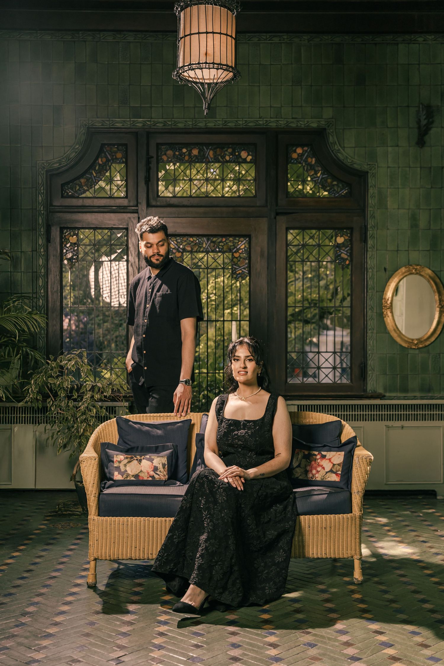 A couple poses on a wicker sofa in a moody, vintage-style interior setting.