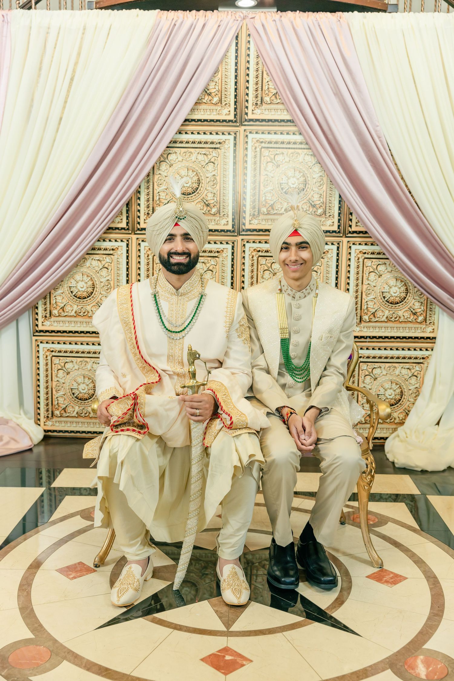 Traditional wedding party members in cream-colored outfits sit on decorated chairs against an elegant backdrop.