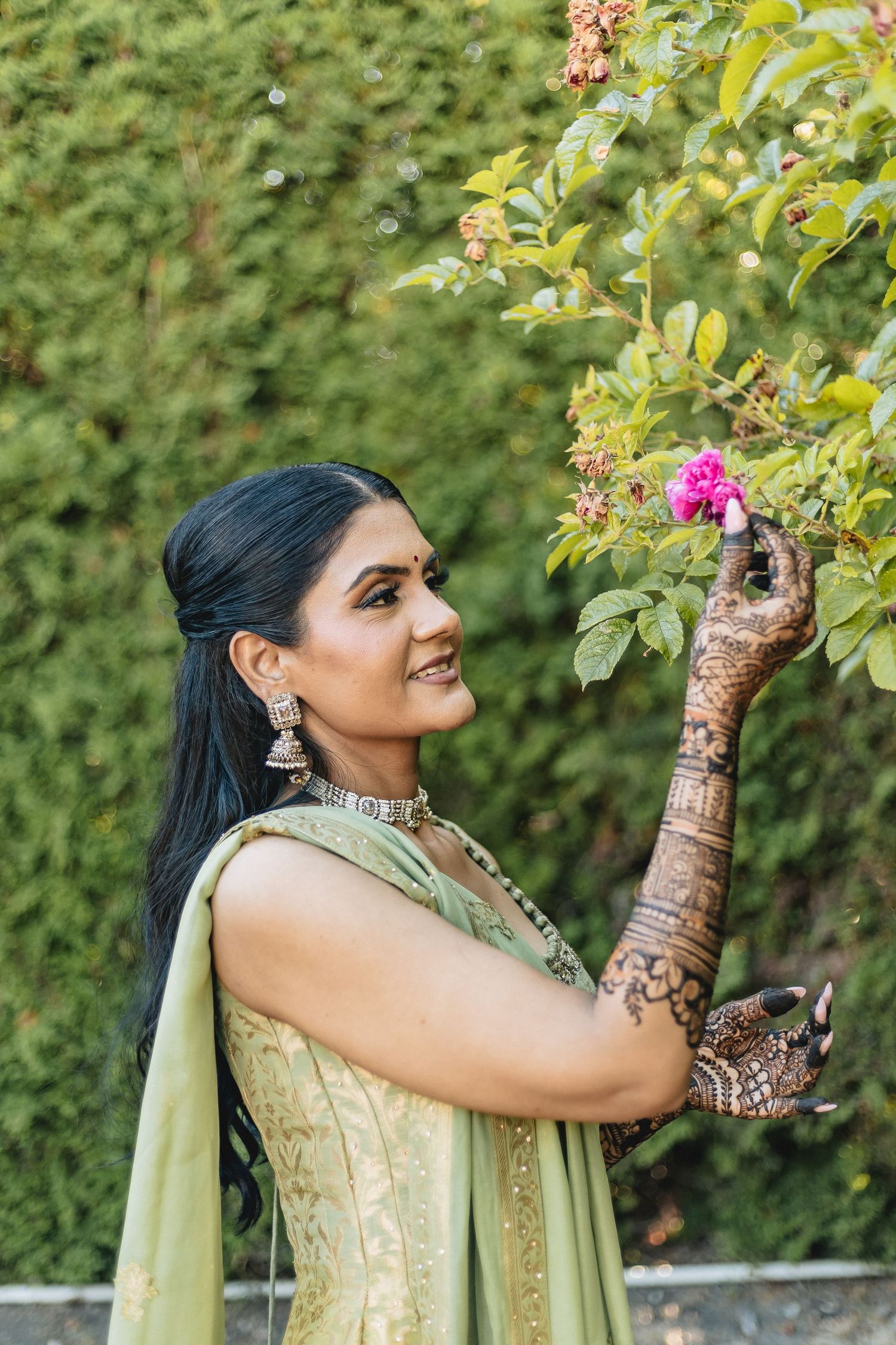 A person in traditional Indian attire with henna-decorated hands reaches towards flowering branches in a garden setting.