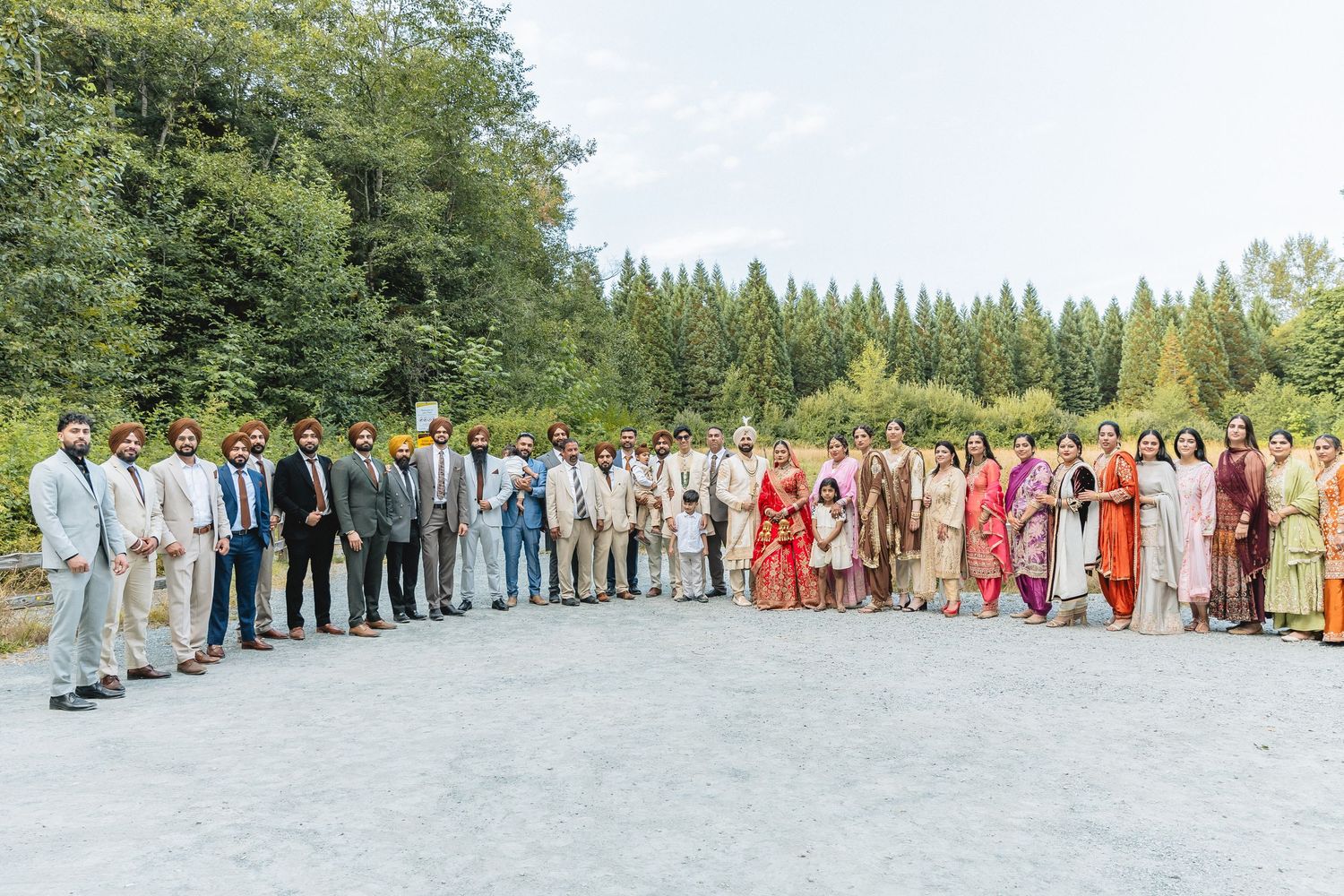 A large group of wedding guests pose together outdoors for a panoramic group photo.
