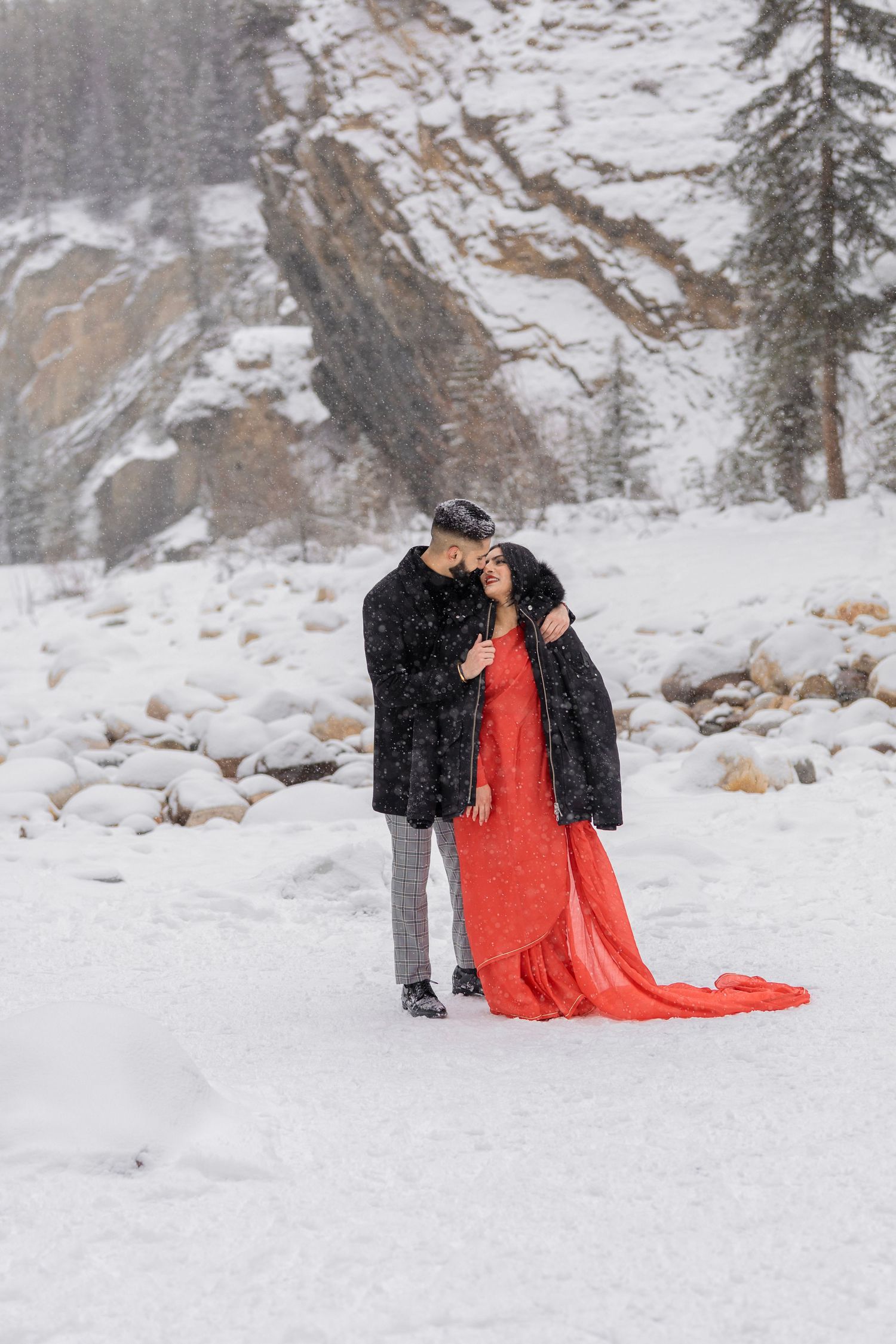 Couple embracing in snowy mountain landscape, woman in red gown and man in black coat, snowfall backdrop.