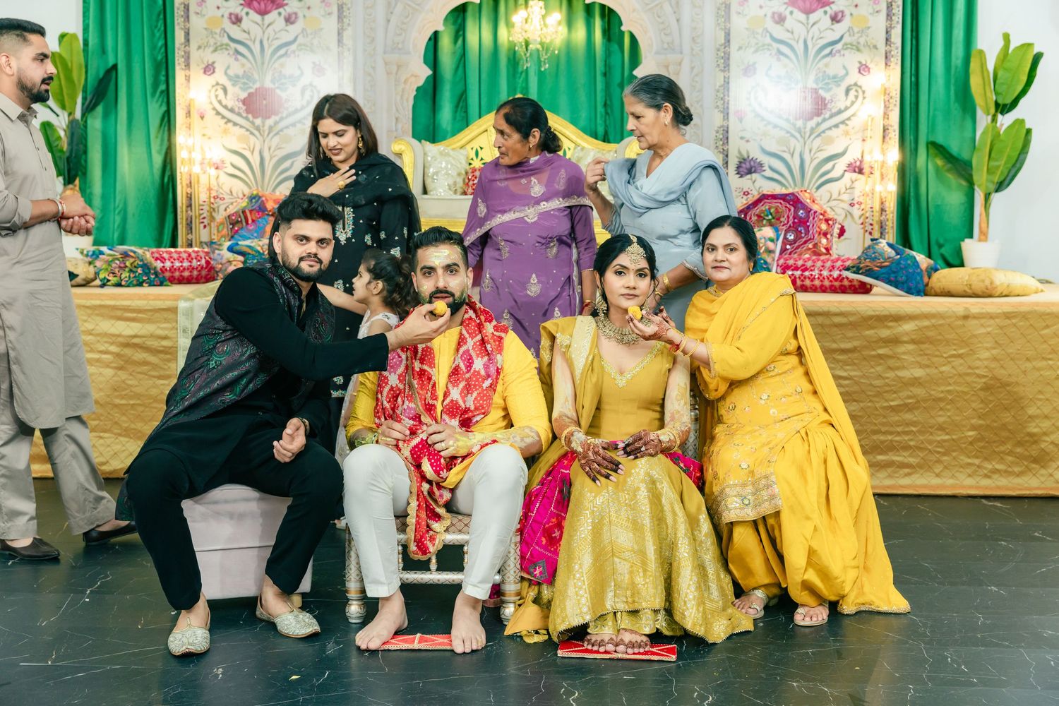 Group photo of wedding party members in colorful traditional Indian attire posing on decorative stage.