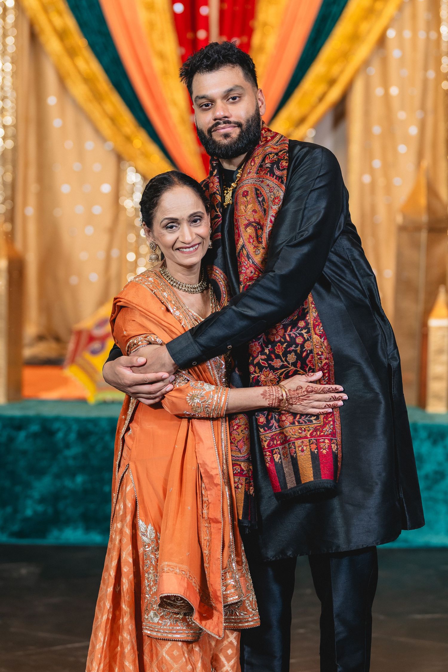 A couple in traditional Indian attire embraces during a wedding celebration with colorful draped backdrop.