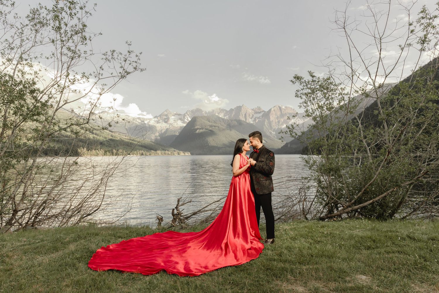 A couple poses dramatically against a mountain lake backdrop with a flowing red dress creating an ethereal effect.