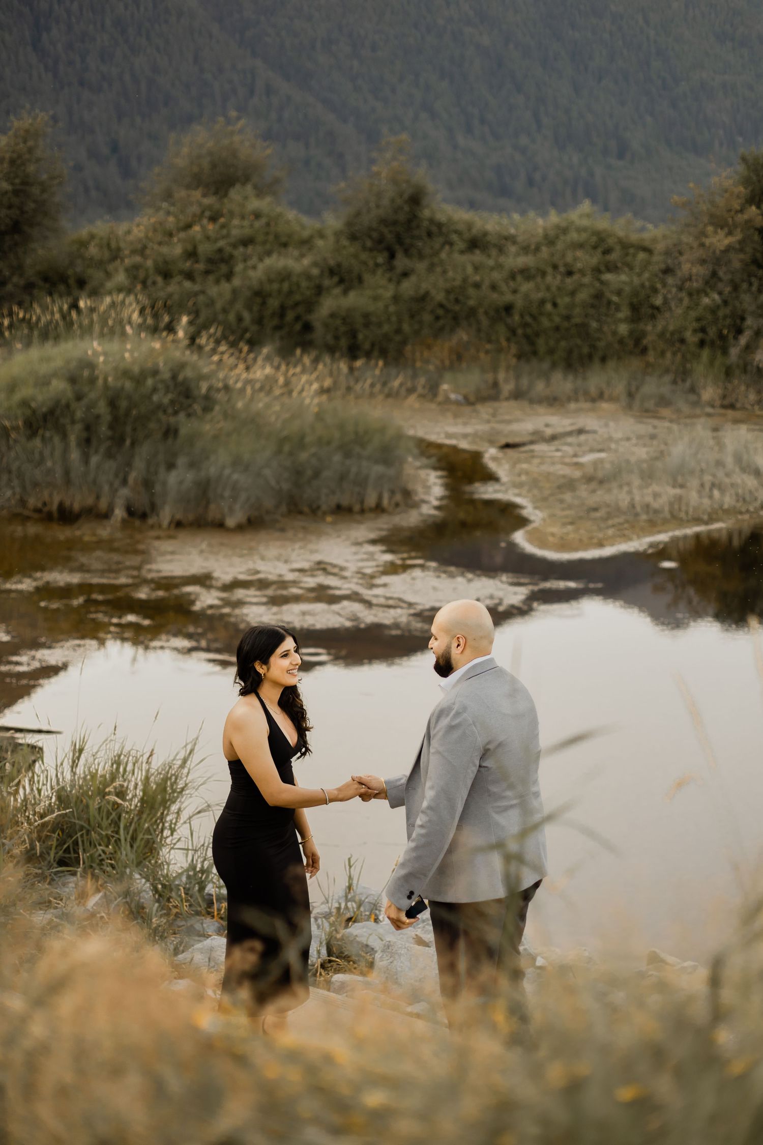 A couple shares a tender moment by a serene lake with mountains in the background during golden hour.
