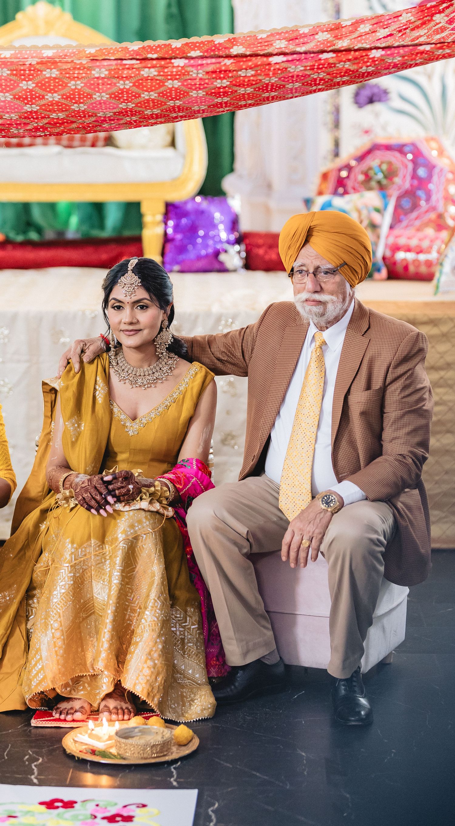 A couple in traditional Indian wedding attire sitting together on a stage with gold and red decorations.