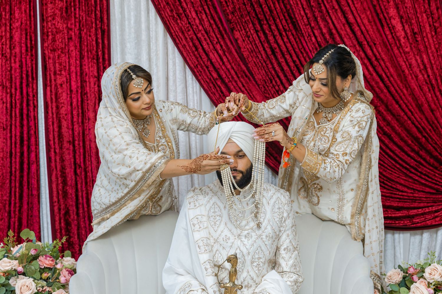 Wedding ceremony participants in traditional white and gold attire against red curtain backdrop.