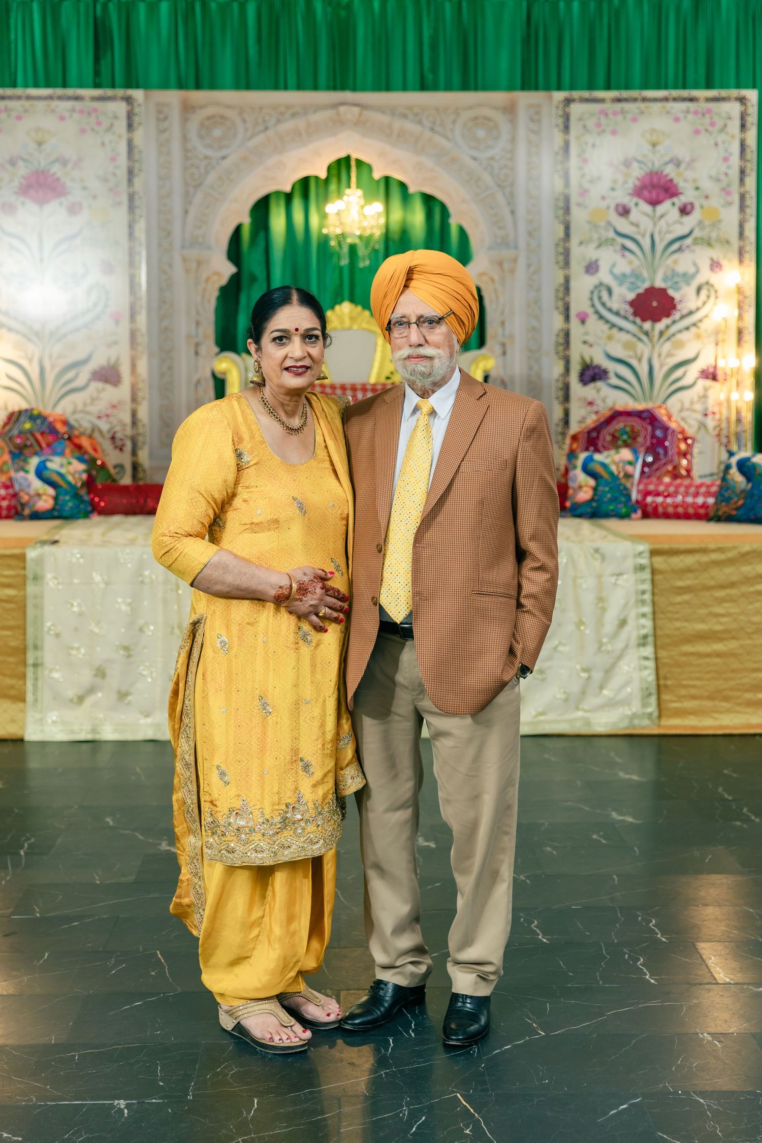 Couple in coordinating yellow and brown formal attire posing in front of decorative green backdrop.