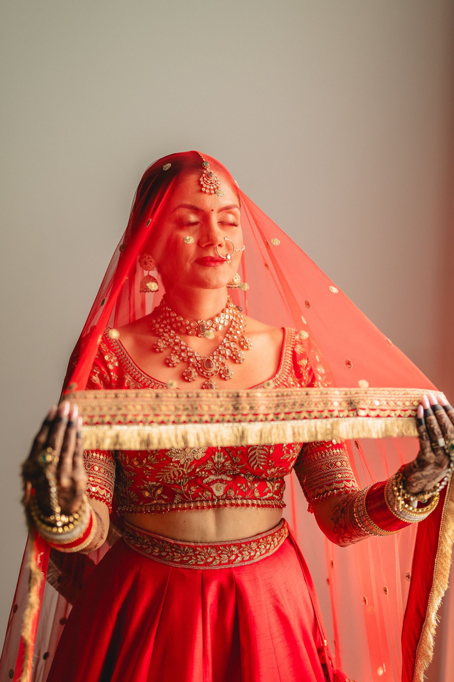 Traditional red bridal lehenga with ornate embroidery and sheer veil in elegant indoor setting.