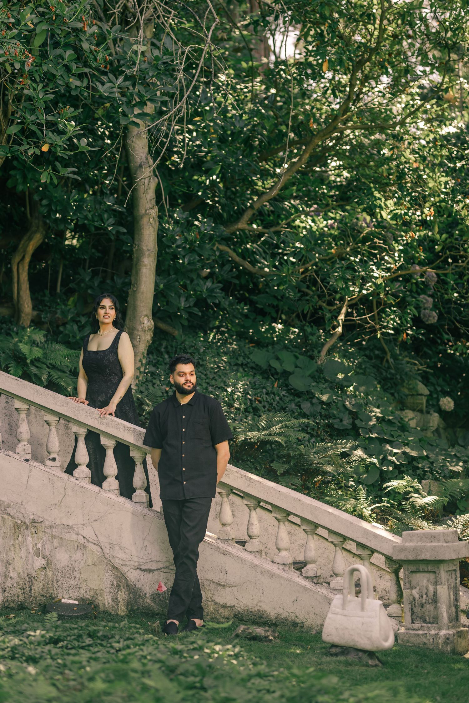 Couple in black formal attire walking along a stone balustrade in a garden setting.