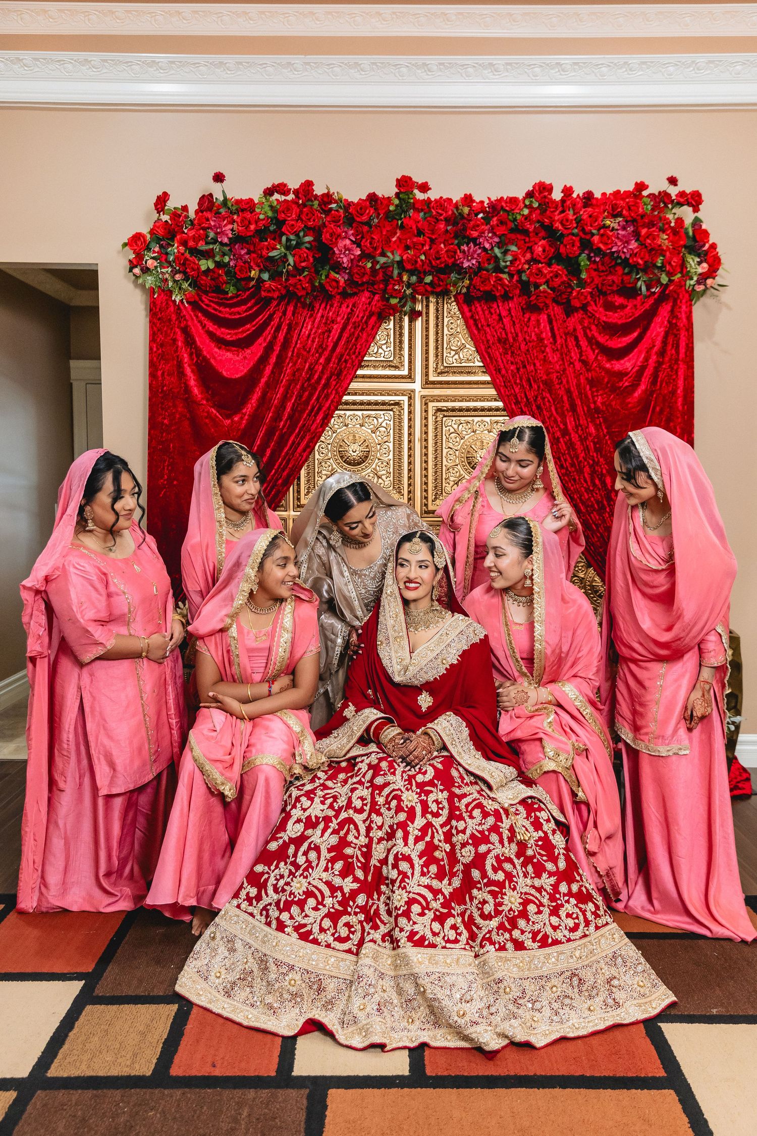 A group in matching pink outfits pose together with a person in an ornate red and gold lehenga for wedding photos.
