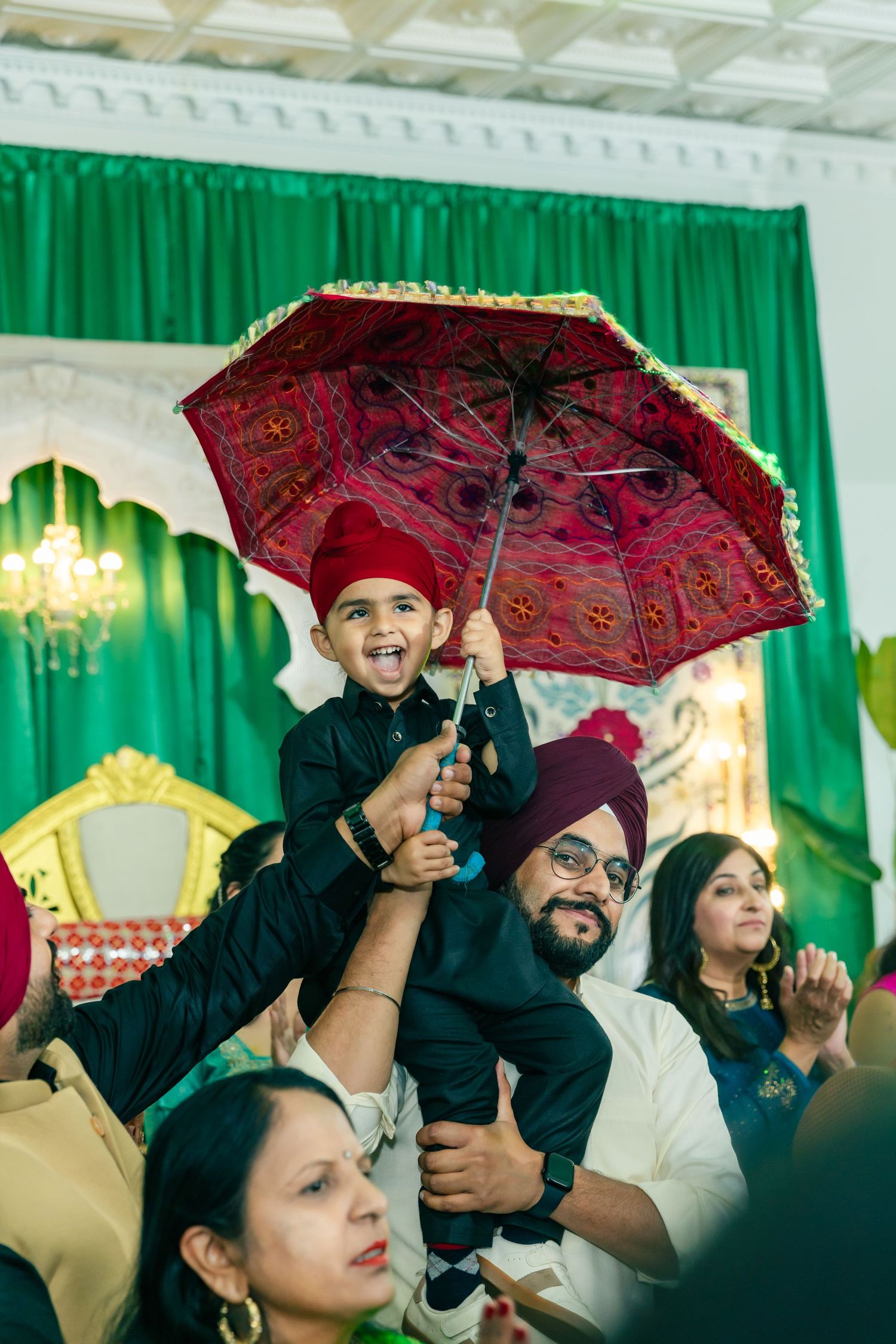 Wedding guests hold decorative red umbrellas during an indoor celebration with green curtains in the background.