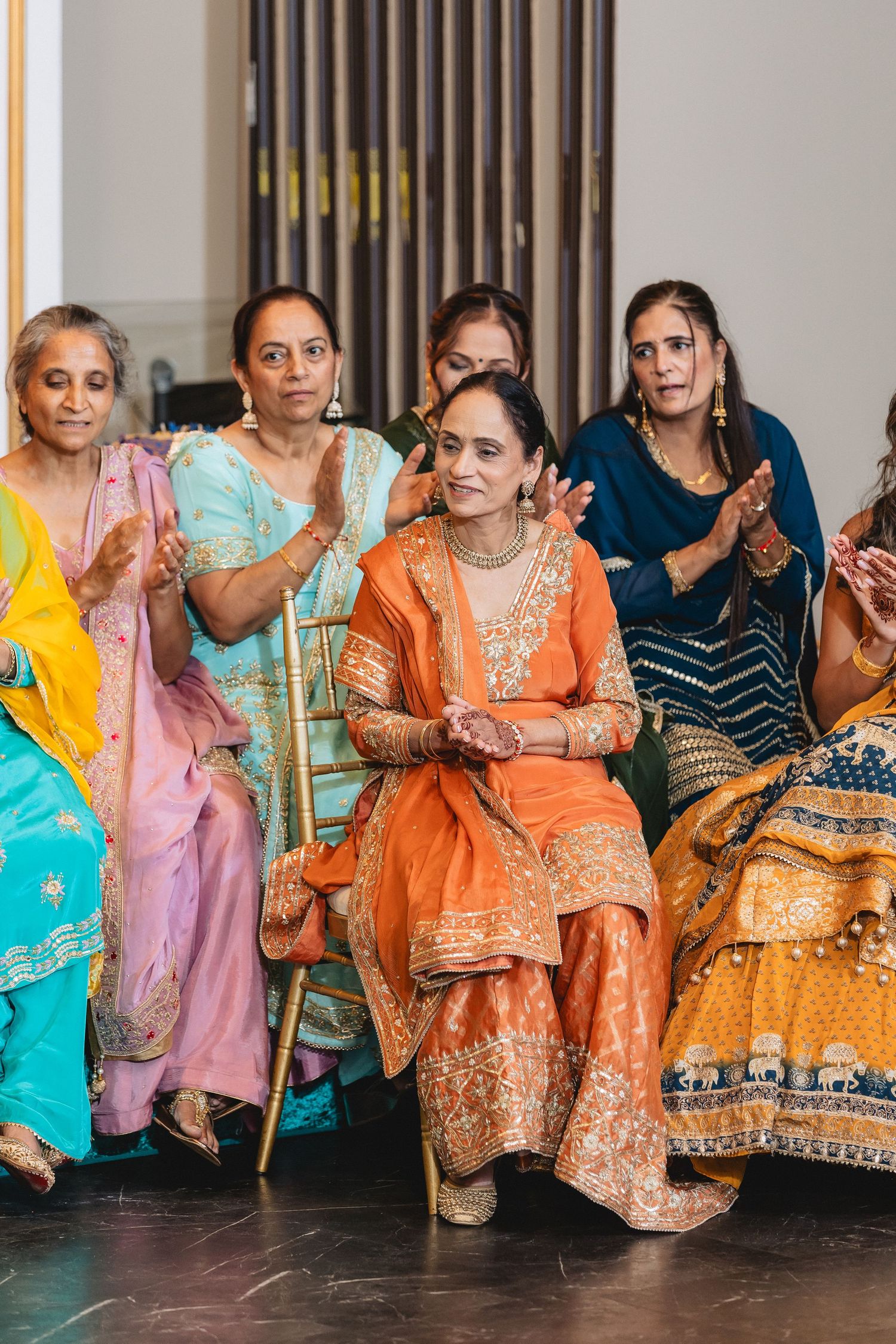 A row of women seated wearing colorful traditional Indian outfits in orange, pink, and turquoise.