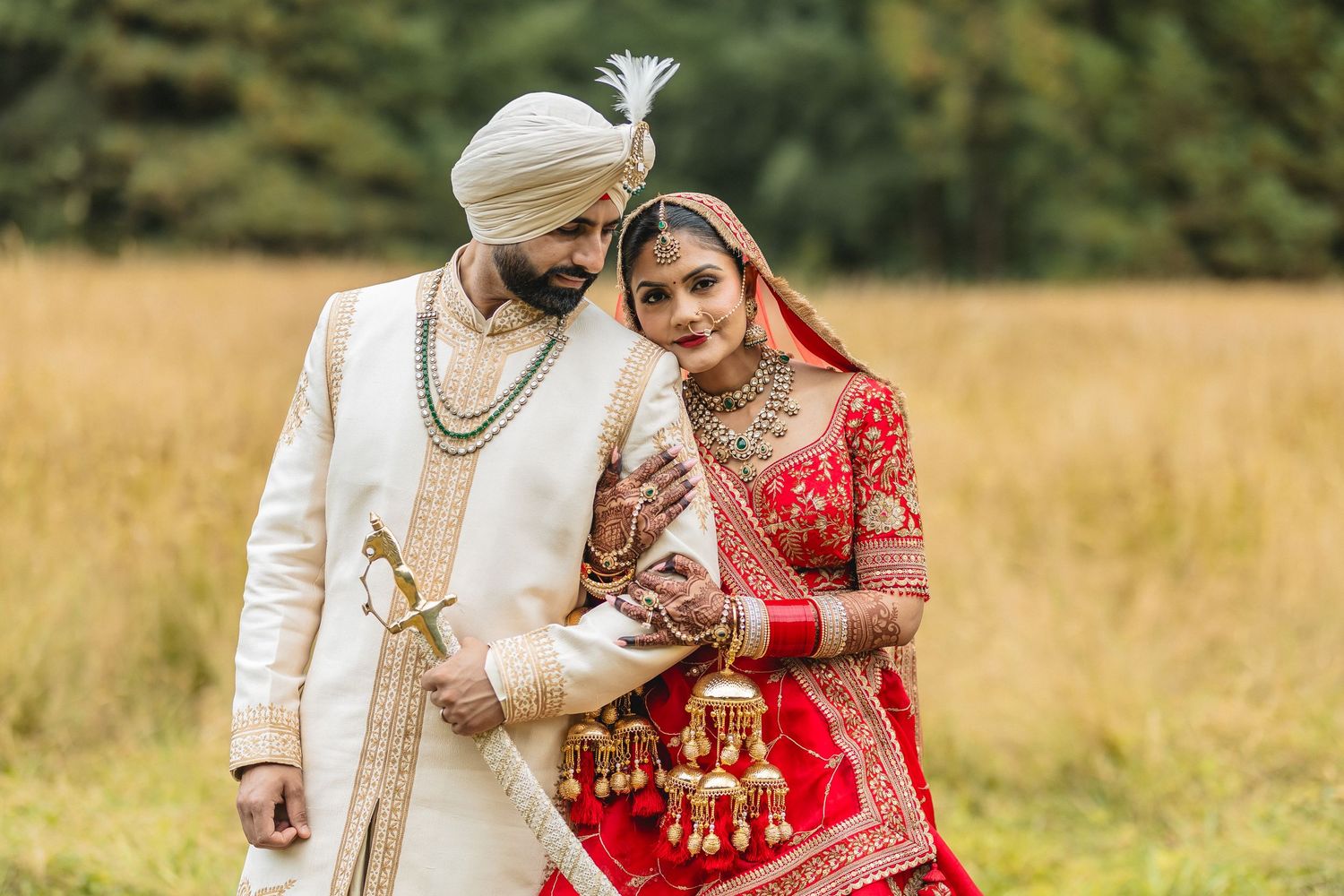 Couple in traditional Indian wedding attire posing in a scenic outdoor field setting.