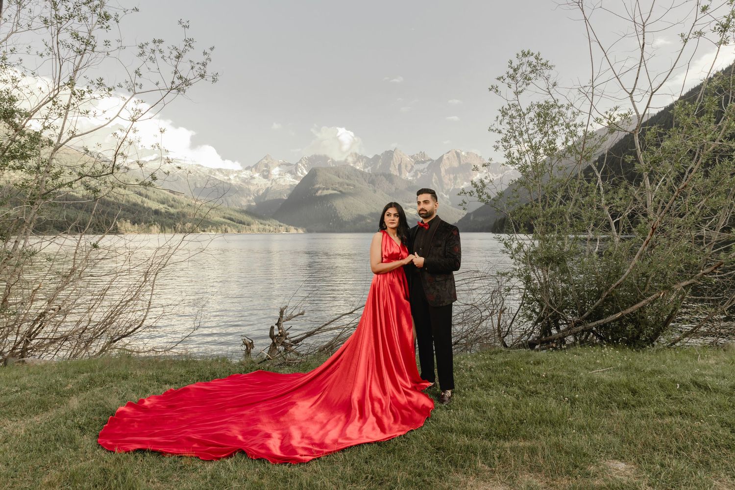 A couple in a red flowing dress poses romantically against a misty mountain lake backdrop at sunset.