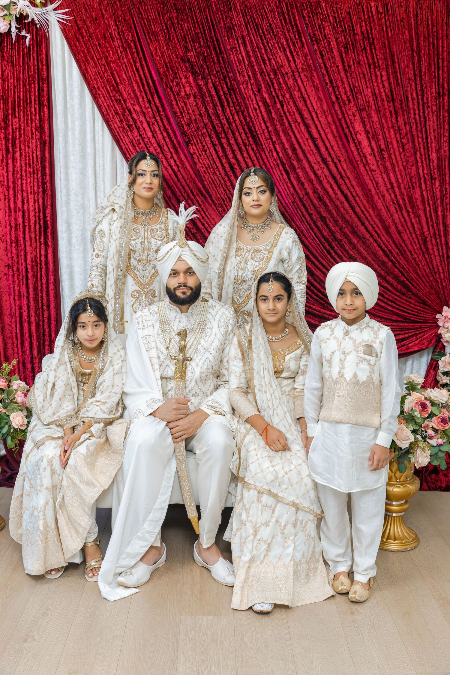 A group in traditional white and gold Indian wedding attire poses against a red curtain backdrop.