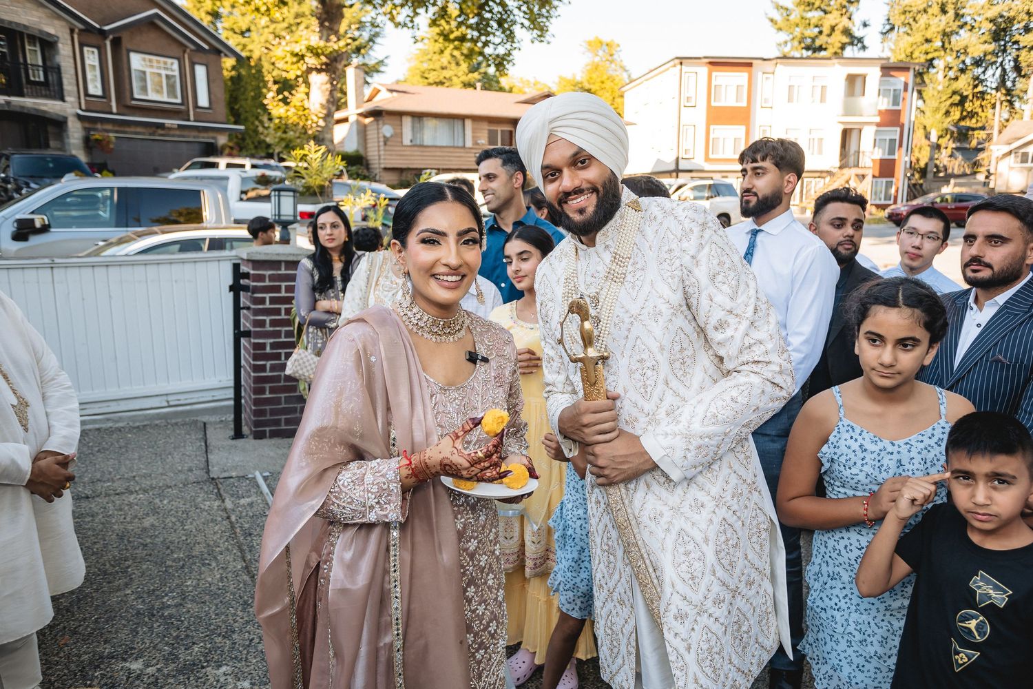 Wedding guests gather outdoors in traditional Indian attire holding celebratory items at a residential venue.