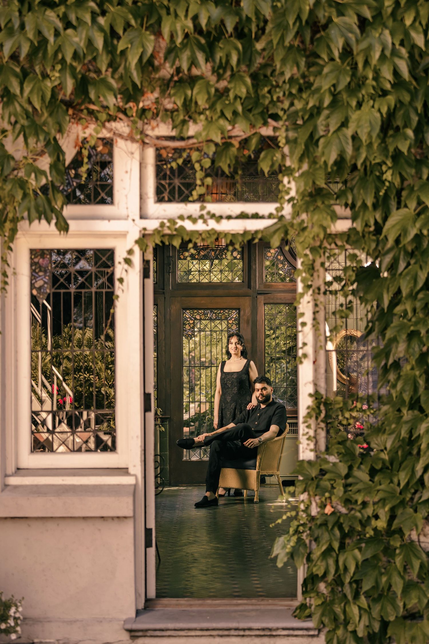 A couple poses in a lush garden setting with white columns and ornate iron gates.