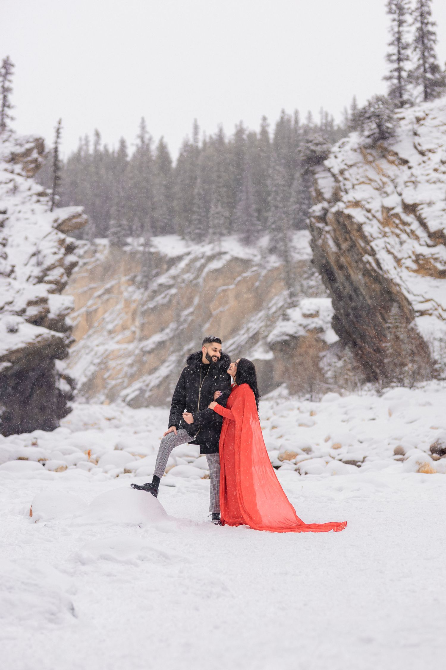 Couple embracing in snowy winter landscape, woman in flowing red dress, man in black jacket, rocky cliffs and pine trees behind.