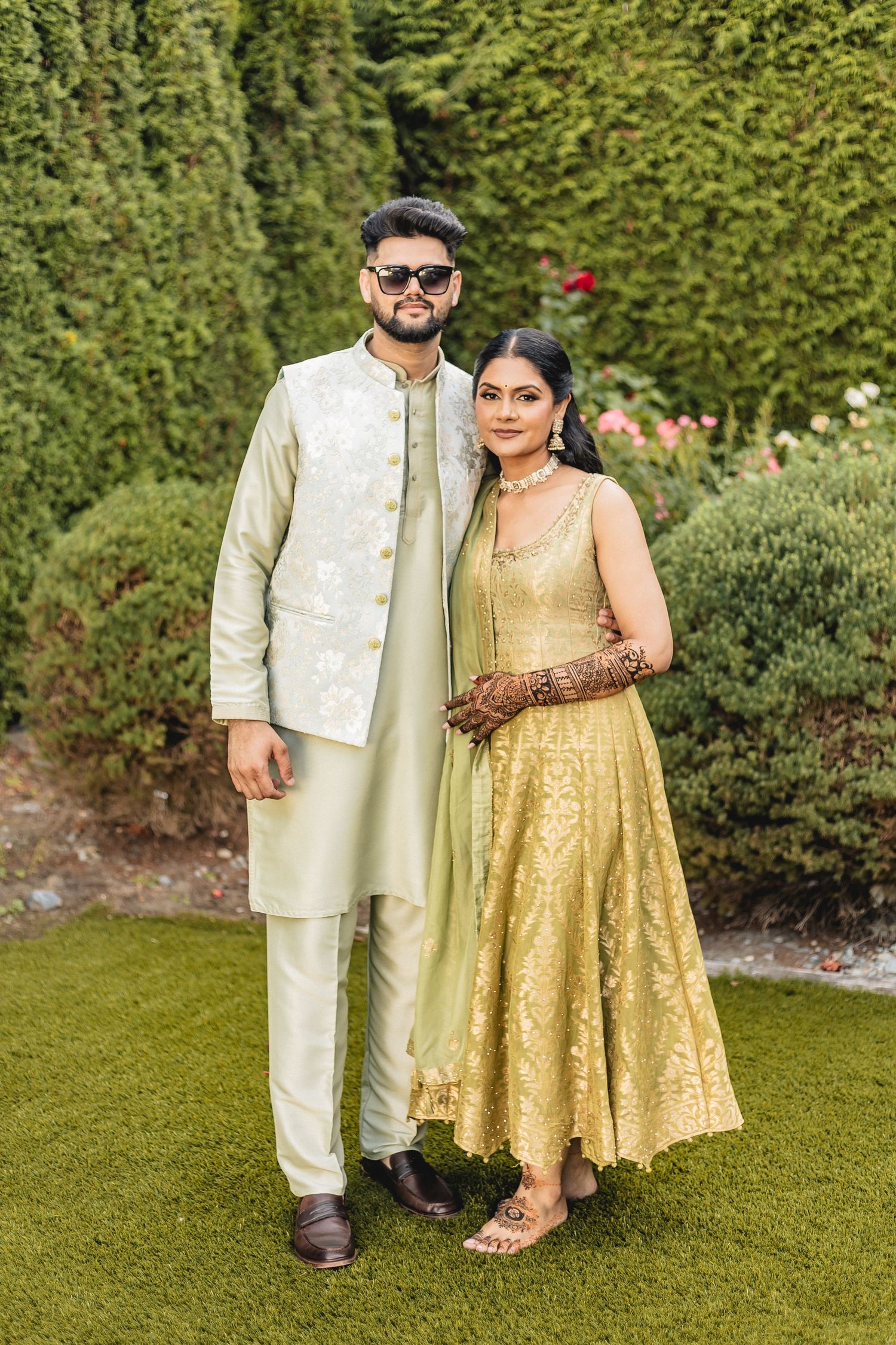 A couple poses outdoors in traditional South Asian attire with gold and cream colored outfits against a lush green garden backdrop.