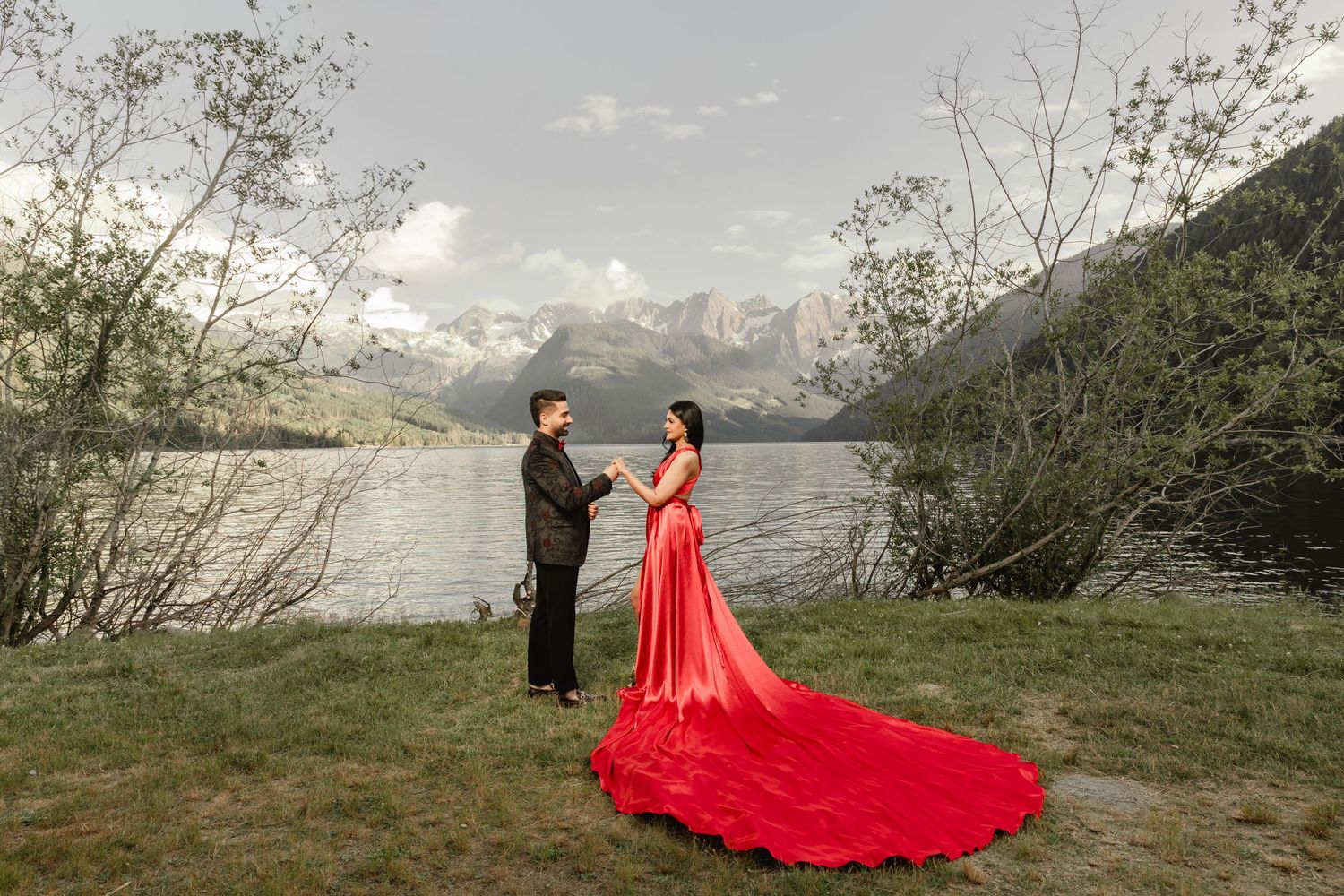 Wedding couple posing by a lake with mountains and dramatic red flowing dress.