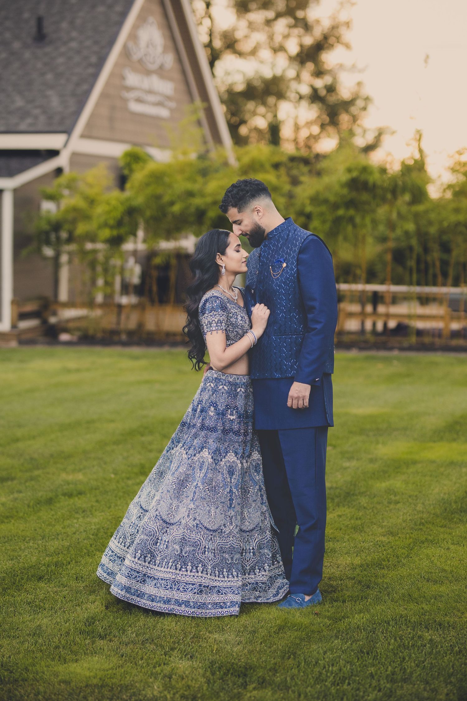 Couple in formal Indian attire poses on grassy lawn during sunset photoshoot.