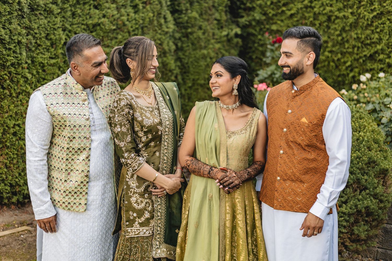 Group wearing traditional South Asian attire poses together in garden with green foliage backdrop.