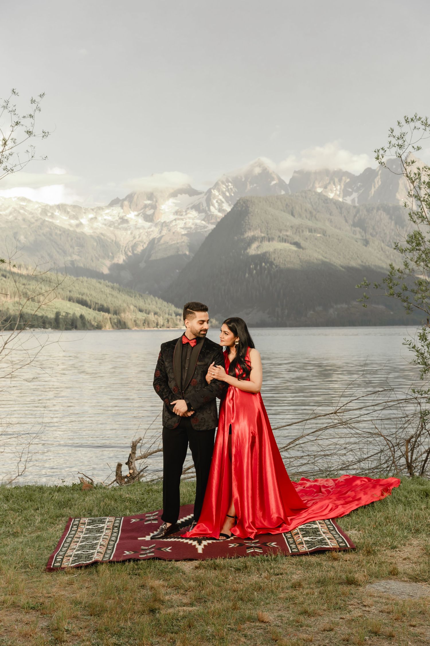 A couple sits on a patterned blanket by a lake with snow-capped mountains in the background.