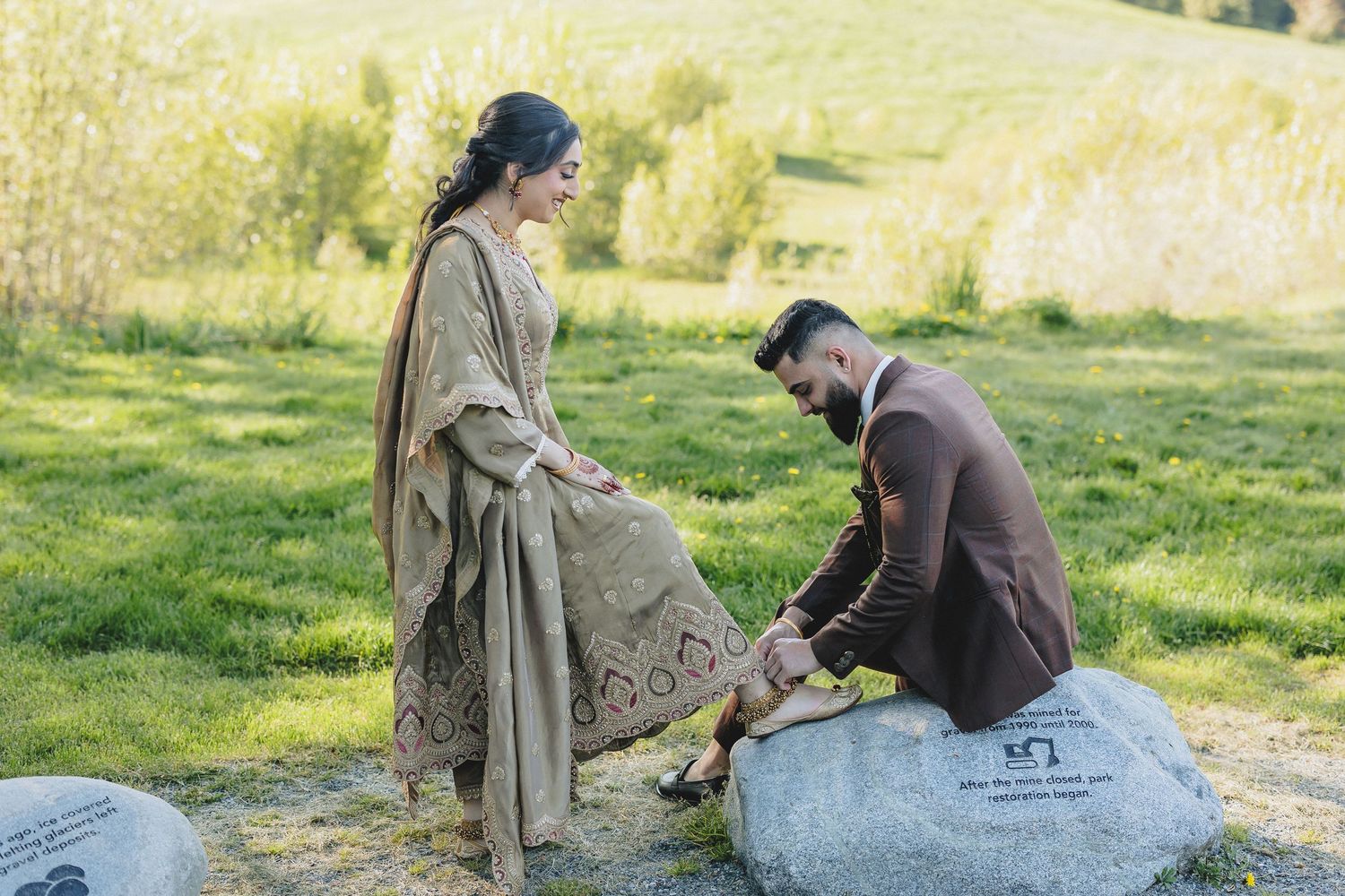 Two people sitting on a large rock in a grassy field during a romantic outdoor moment.