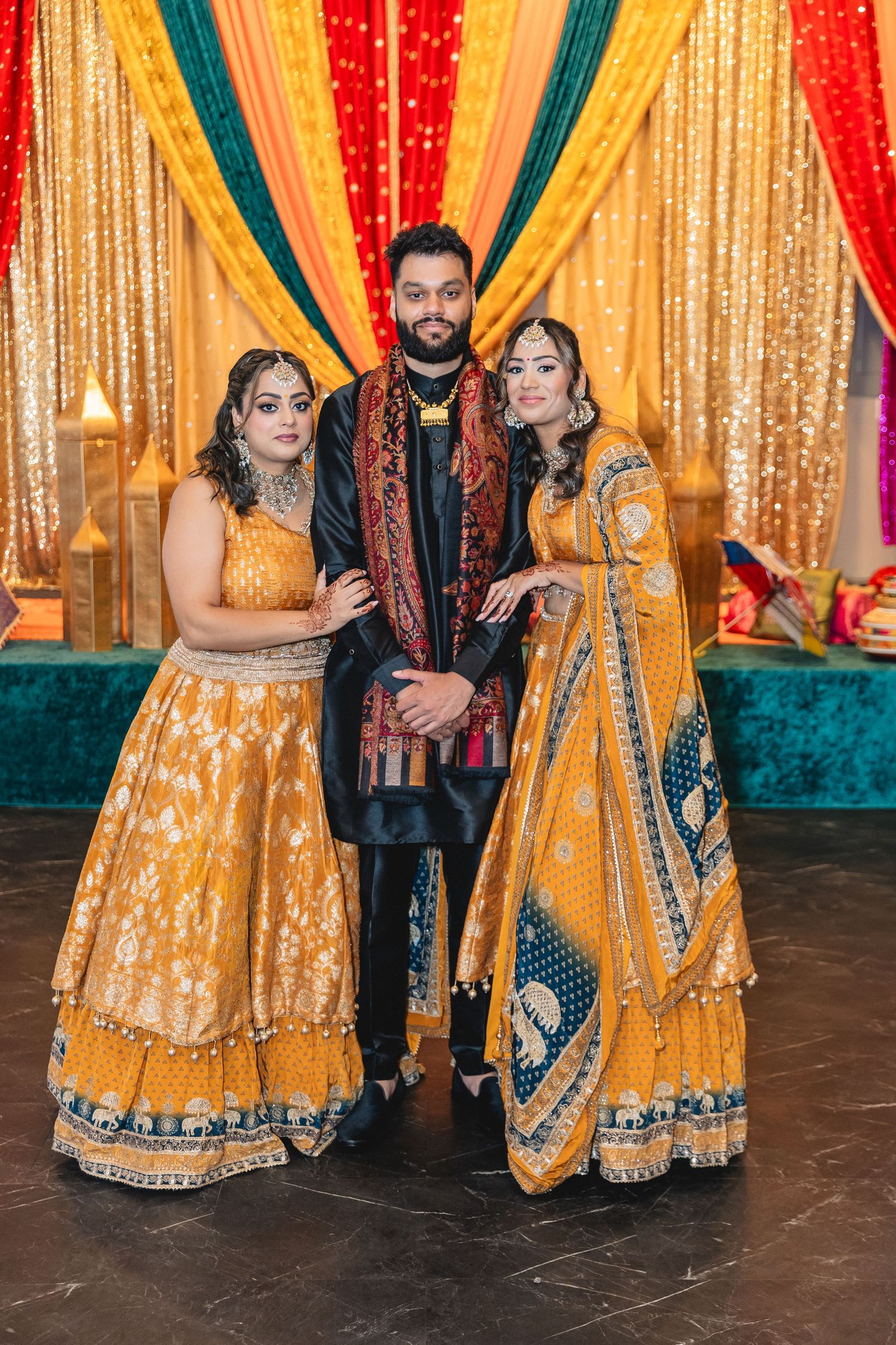 Guests in traditional Indian wedding attire pose against a gold and red backdrop with elegant decorations.