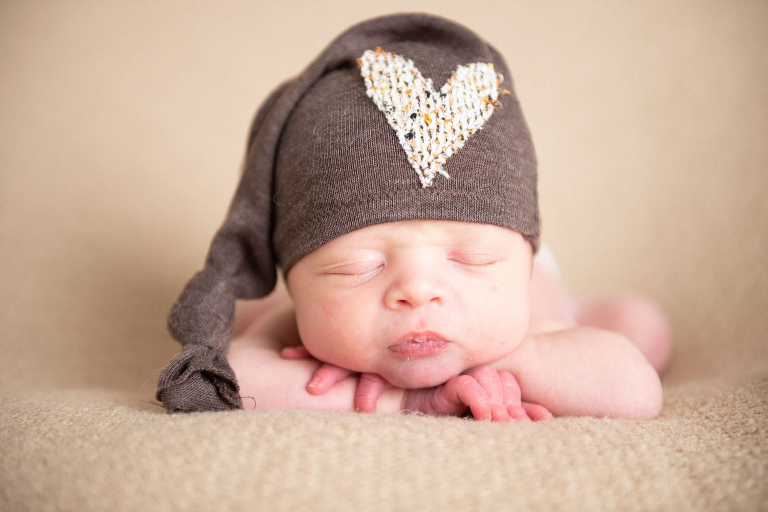 Newborn baby sleeping peacefully in brown knit hat on beige backdrop.