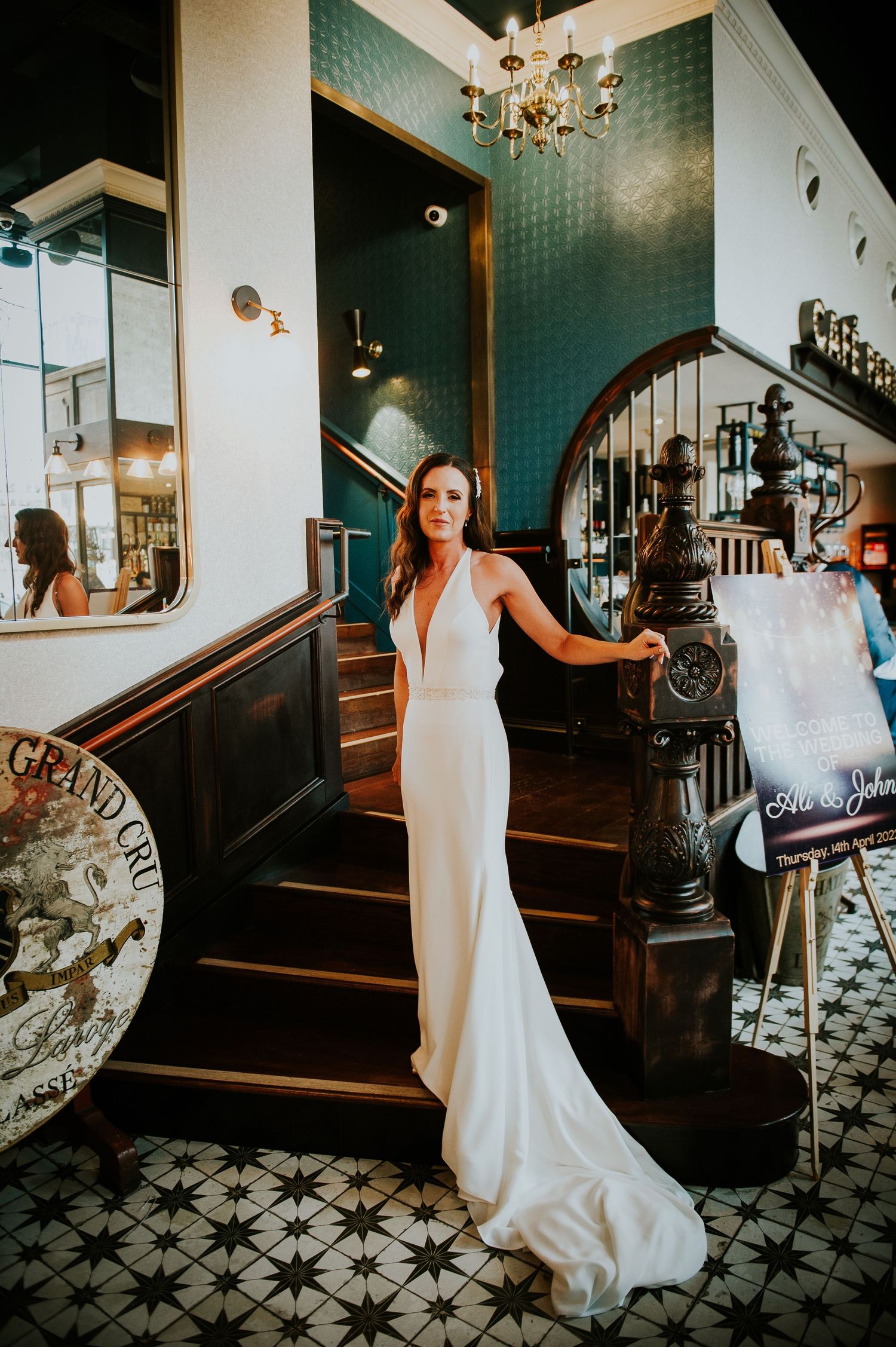 Elegant bride in white gown descending ornate staircase with patterned floor and chandelier in vintage venue.