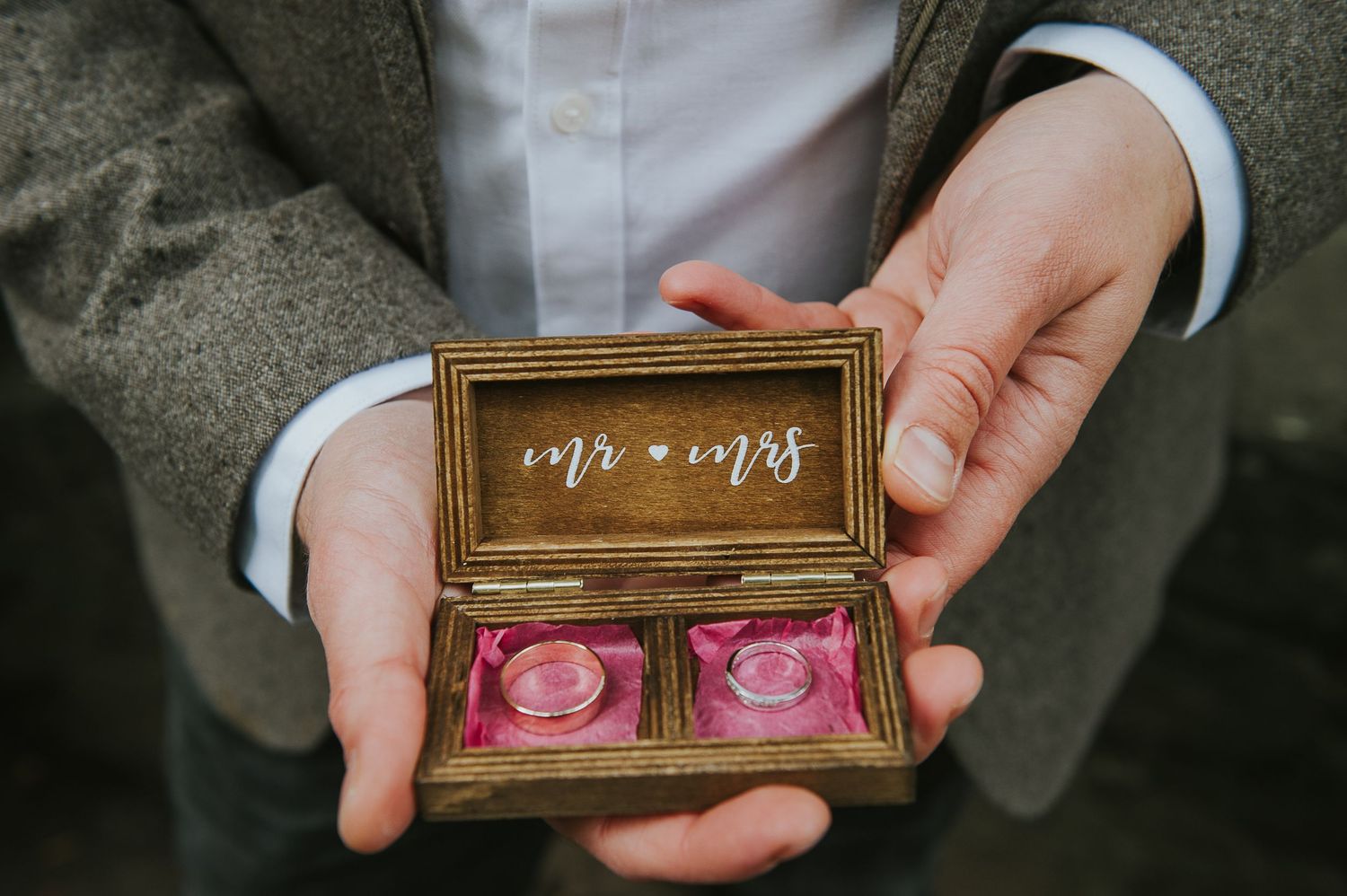Hands in formal attire hold an ornate wooden box containing two pink rose-shaped wedding rings.