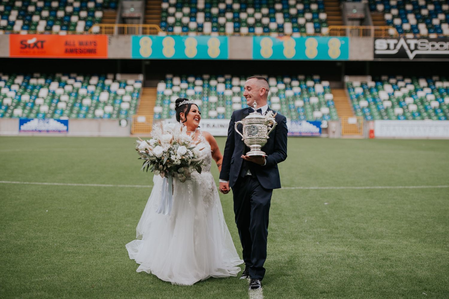A couple walks together on a green sports field stadium during their wedding celebration.
