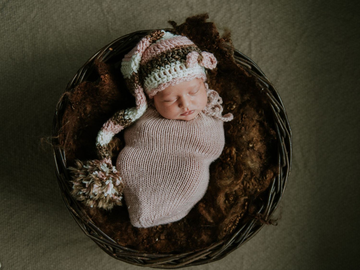 Baby resting in rustic wooden bowl wearing cozy knit wrap against dark background.
