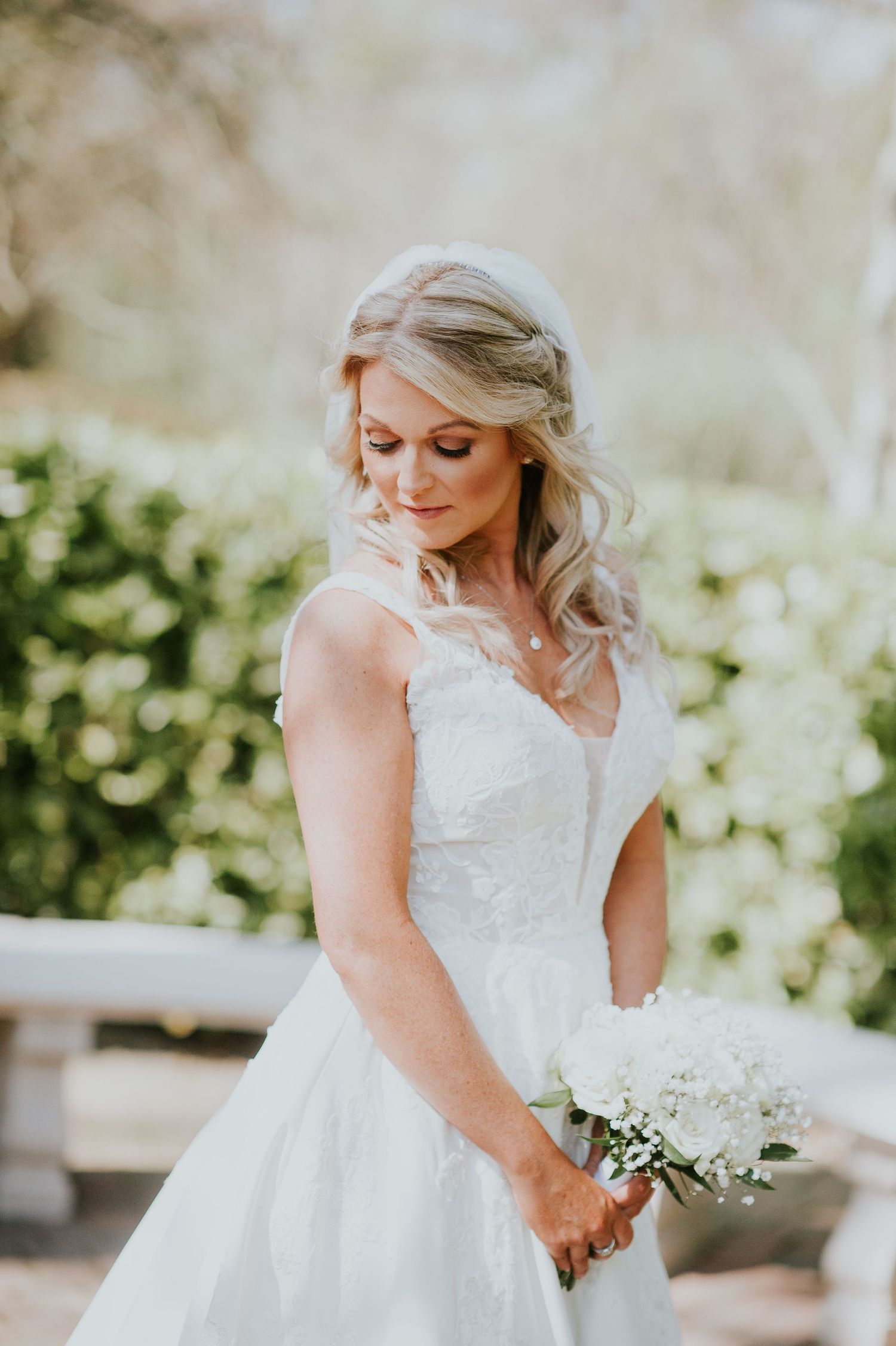 Bride in classic white ballgown poses with bouquet in sunlit outdoor setting.