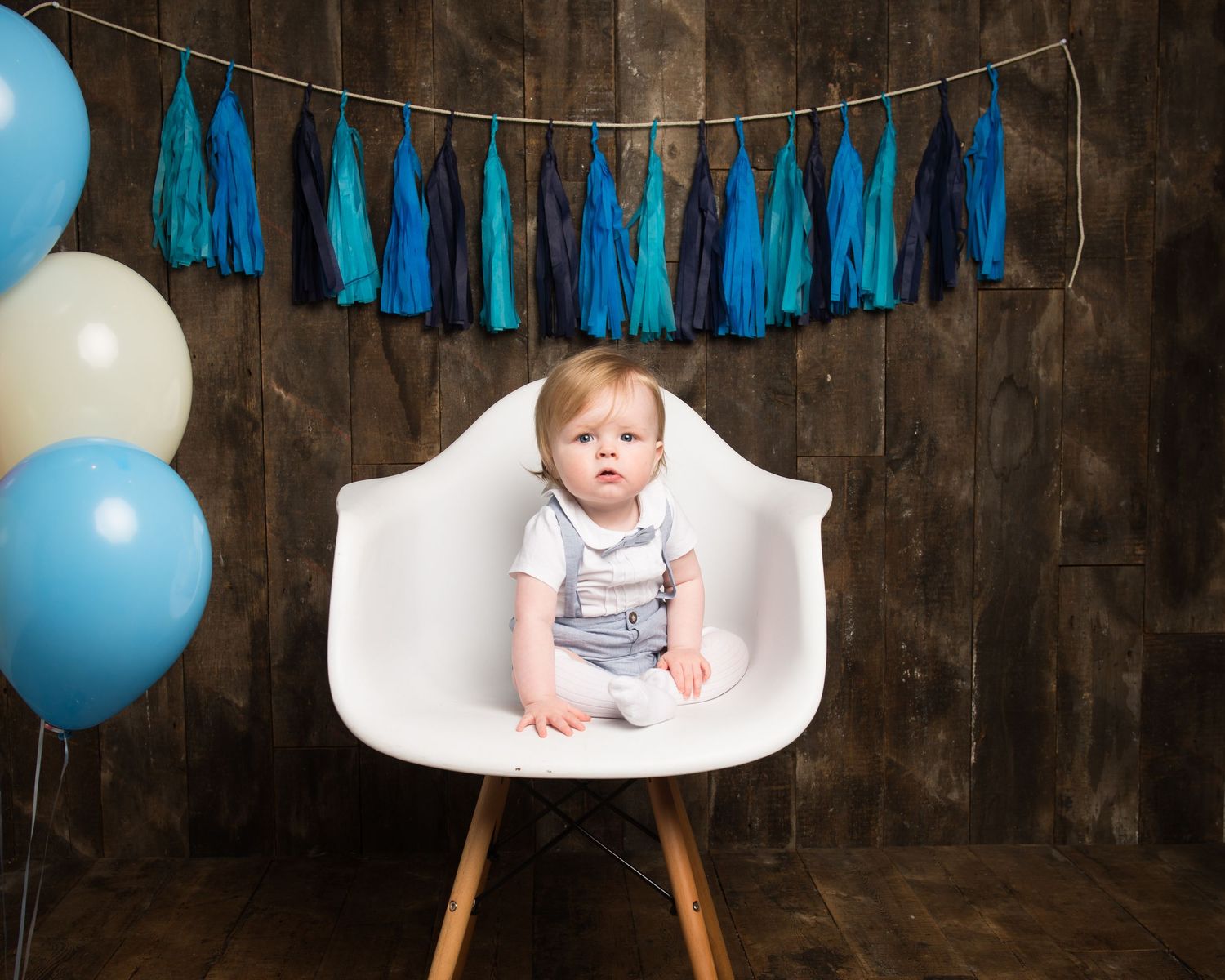 Baby sitting in white modern chair with blue balloon and tassel decorations.