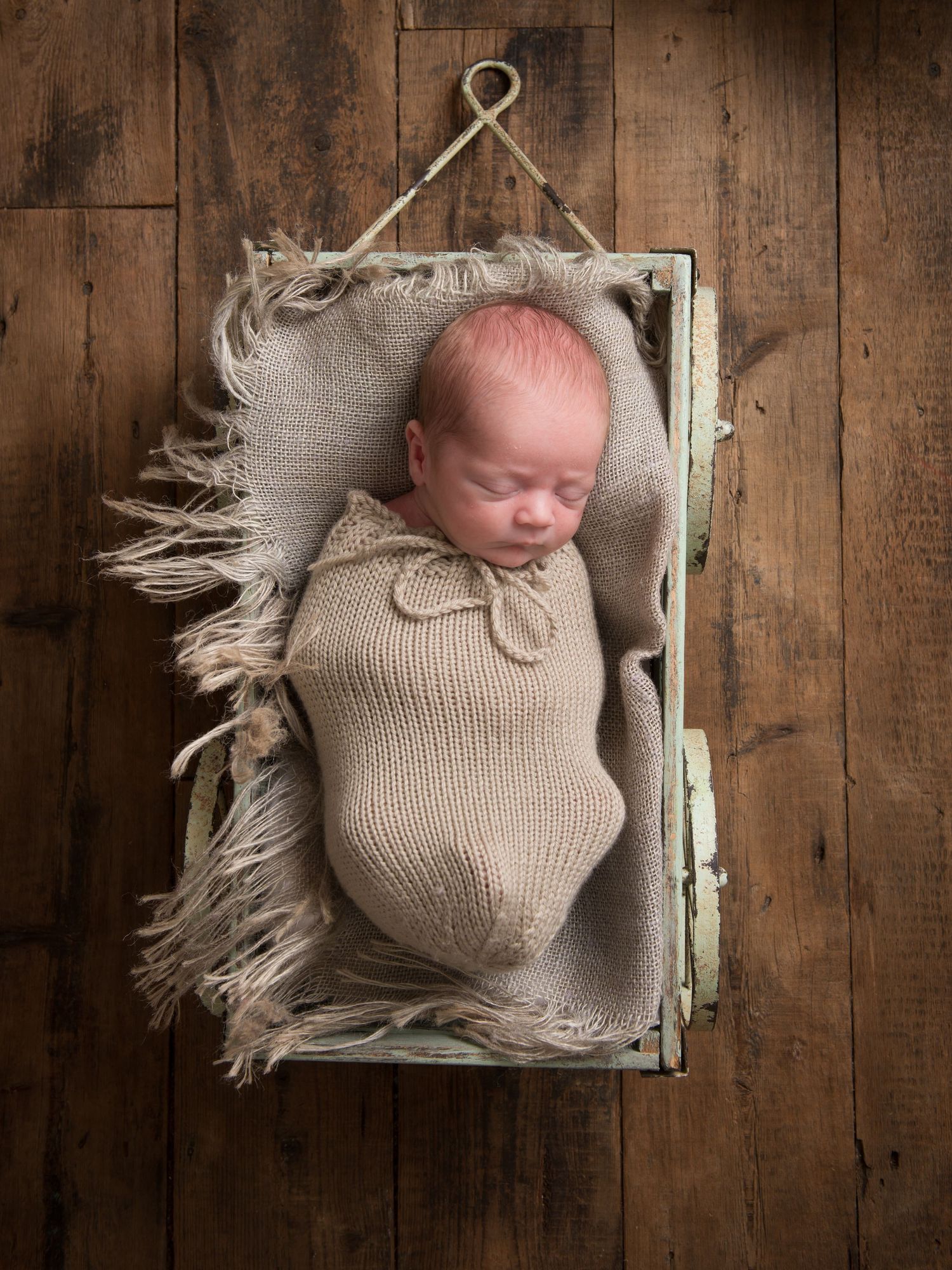 Newborn baby wrapped in beige knit swaddle sleeping peacefully in rustic wooden box with textured fabric.