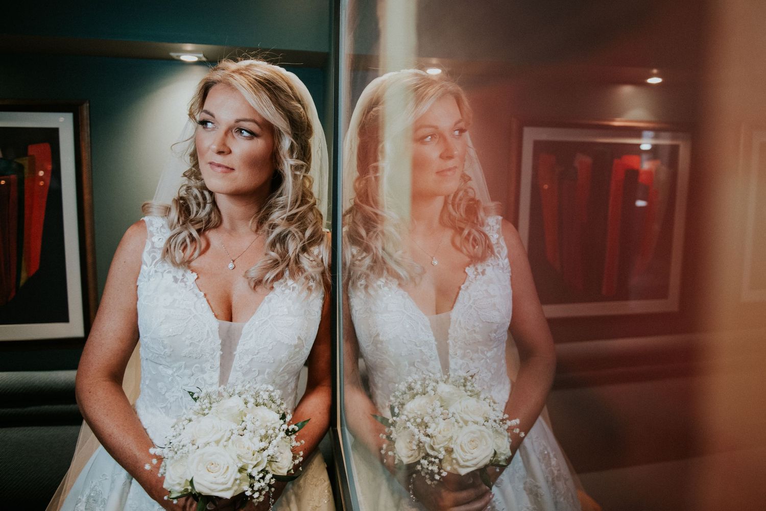 Bride in lace wedding dress poses with bouquet reflected in mirror.