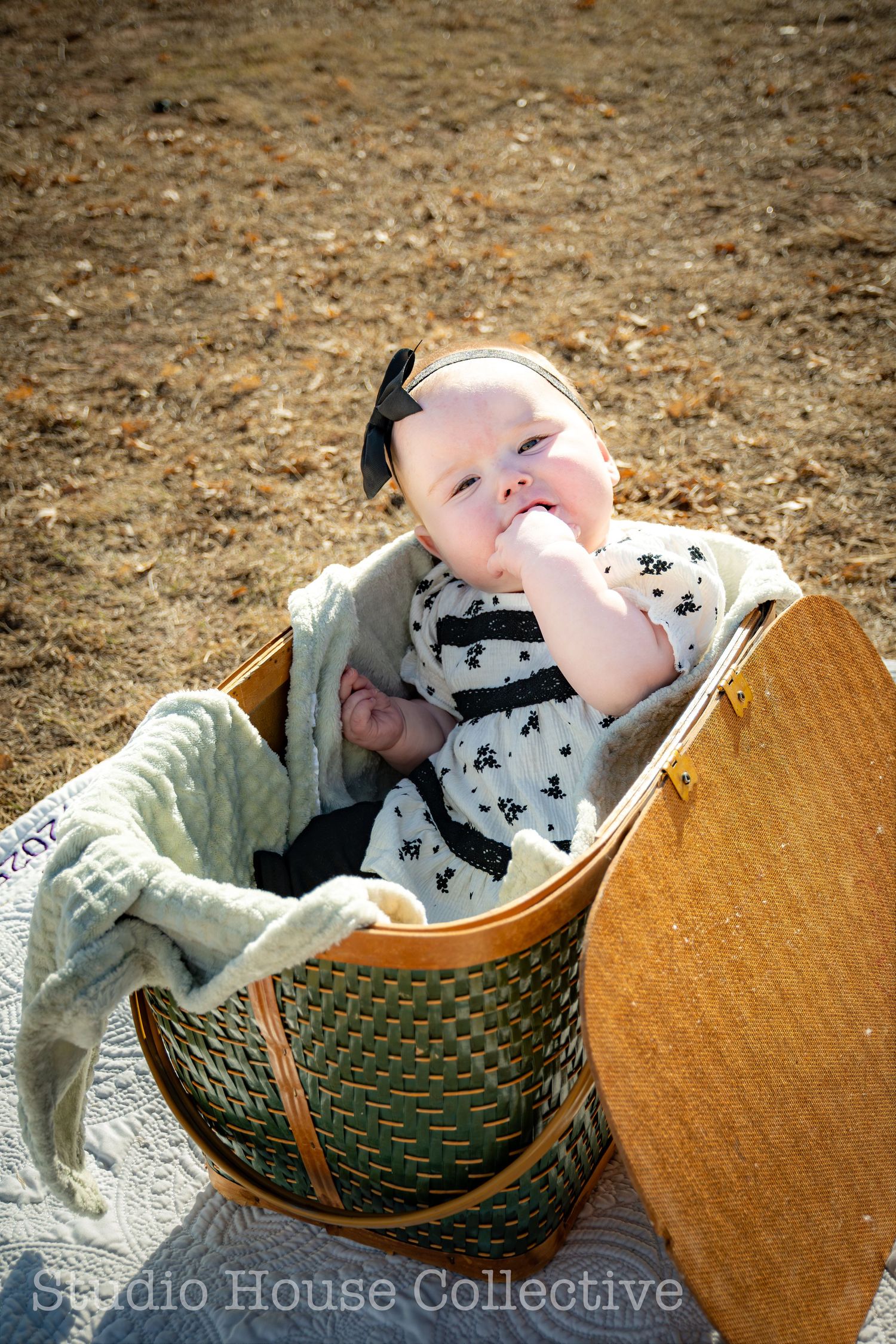 Small baby wearing a black and white patterned outfit sits in a woven basket with a blanket on brown ground.