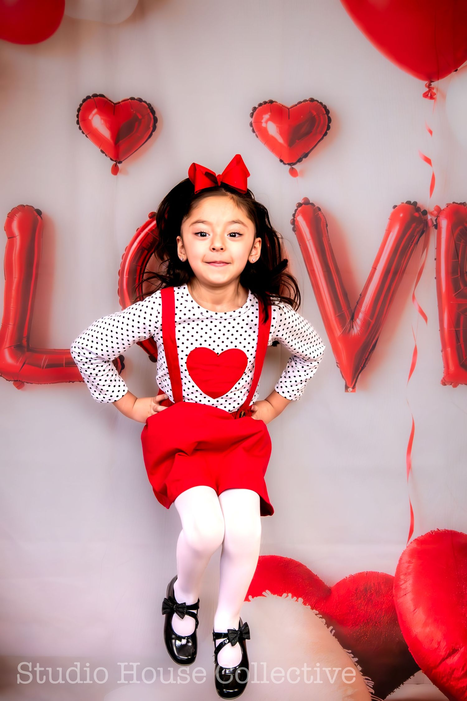 A child in a Valentine's themed outfit with polka dots, red suspenders and bow poses in front of heart balloons and LOVE letters.