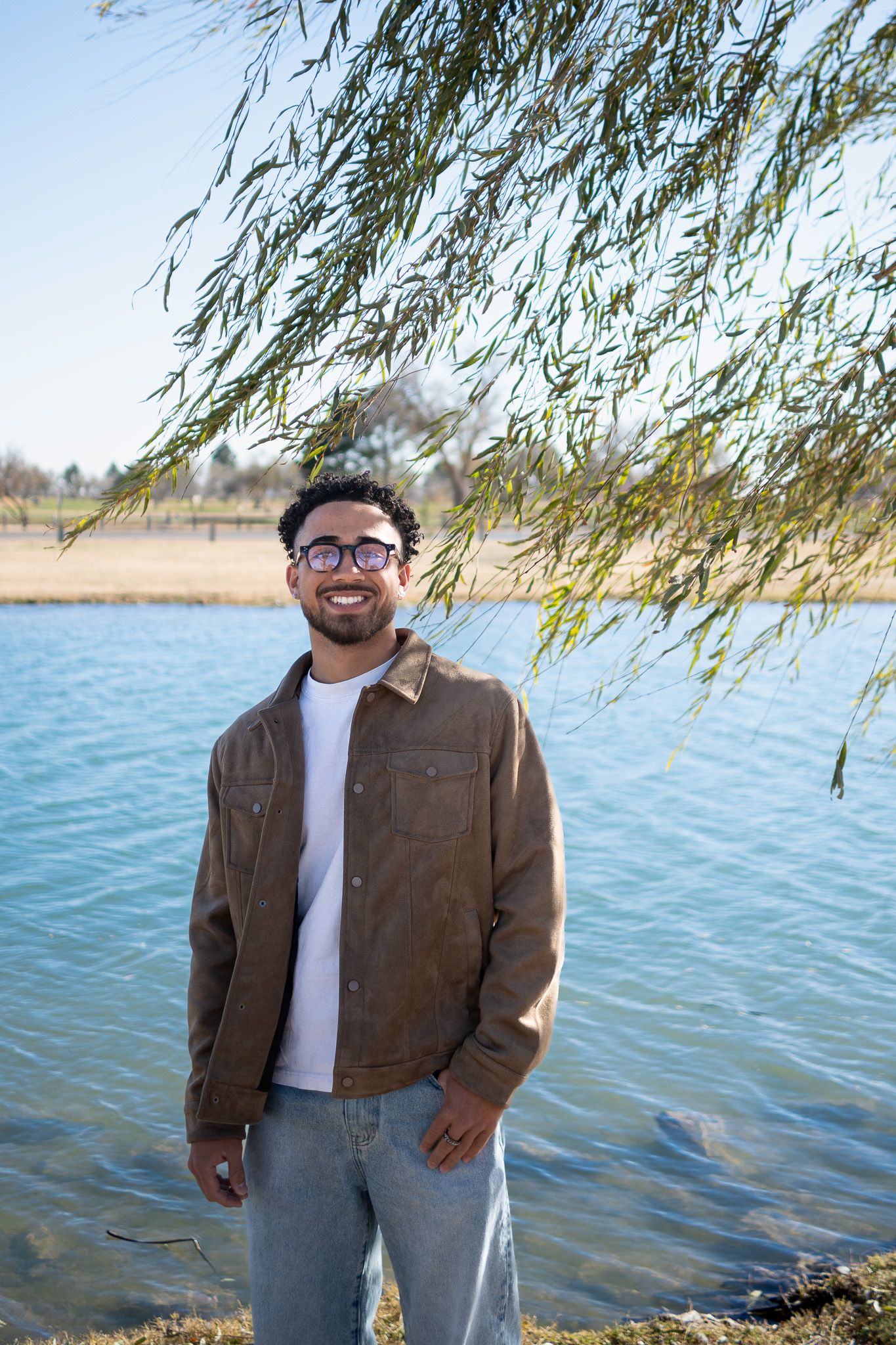Person in brown jacket and white shirt smiling by a lake on a sunny day with willow branches overhead.
