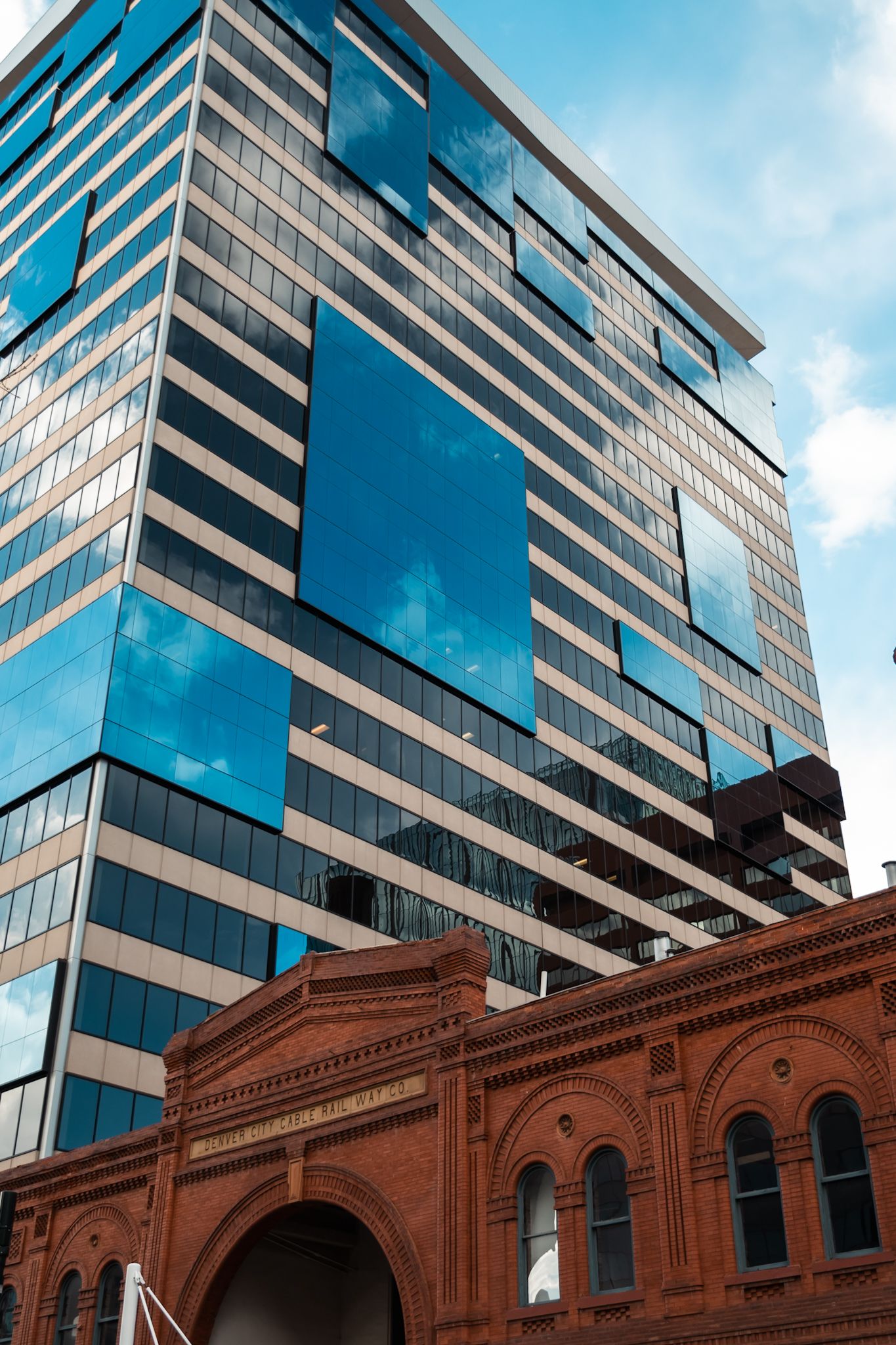 Modern glass office tower rises above historic red brick arched building creating striking architectural contrast against blue sky.