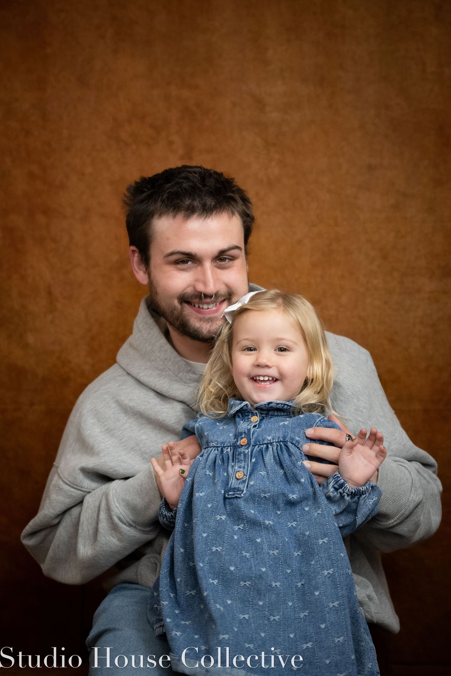 Studio portrait of a parent and young child in denim overalls against a brown background.
