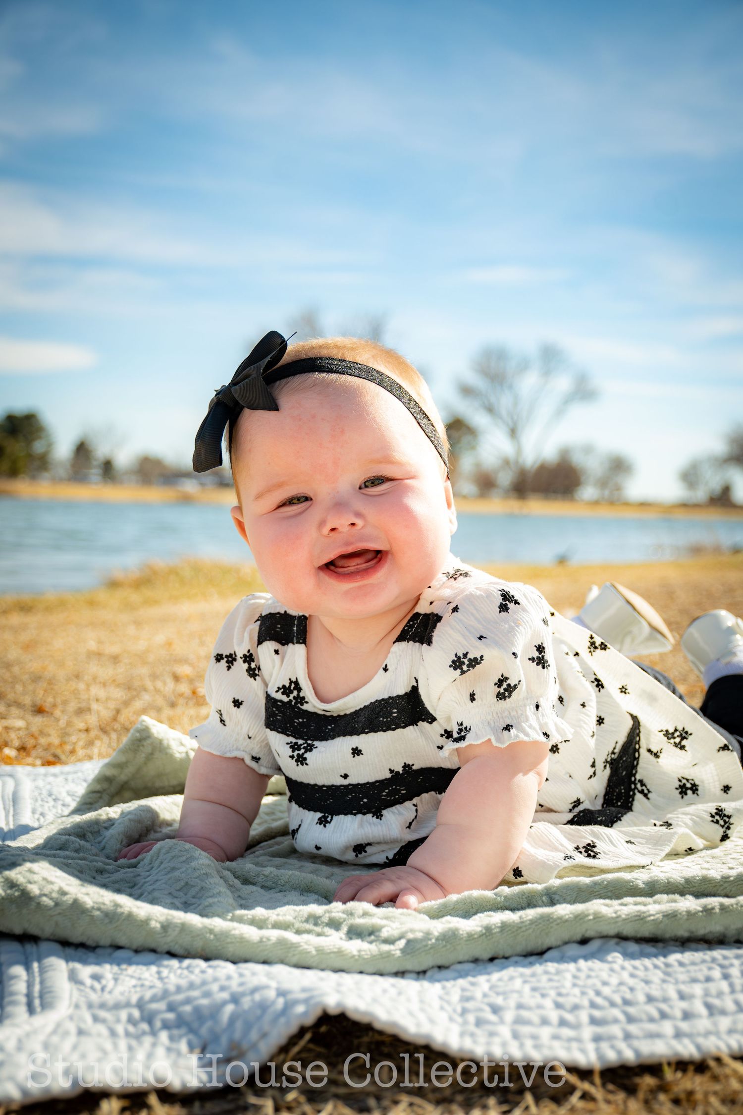 Baby in black and white patterned dress with bow headband smiles while lying on blanket near lake on sunny day.